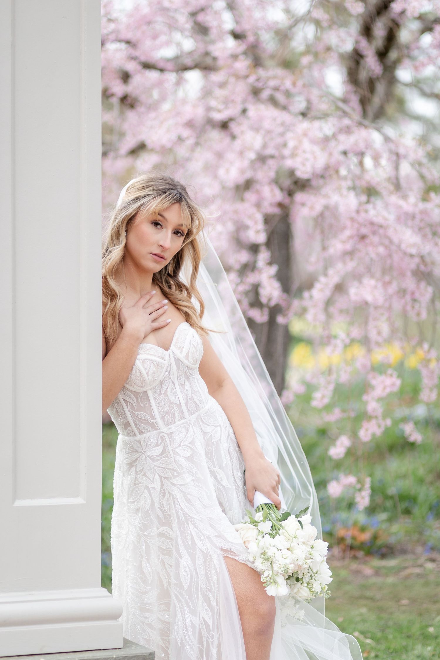 Bride in lace gown with veil poses by column near pink cherry blossom tree.