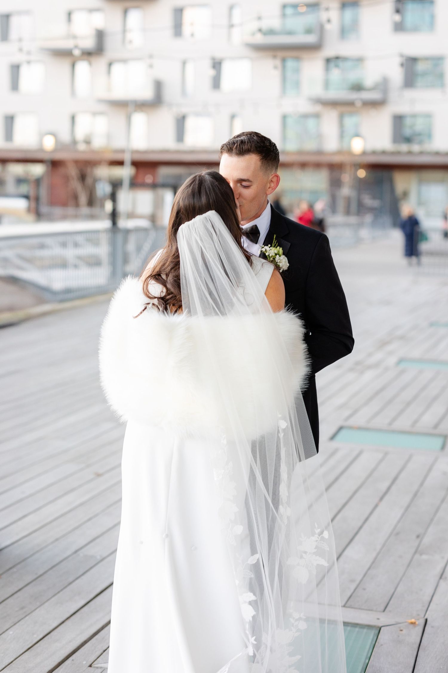 A bride in a white gown with fur wrap and veil shares a kiss with a groom in a black tuxedo outdoors.