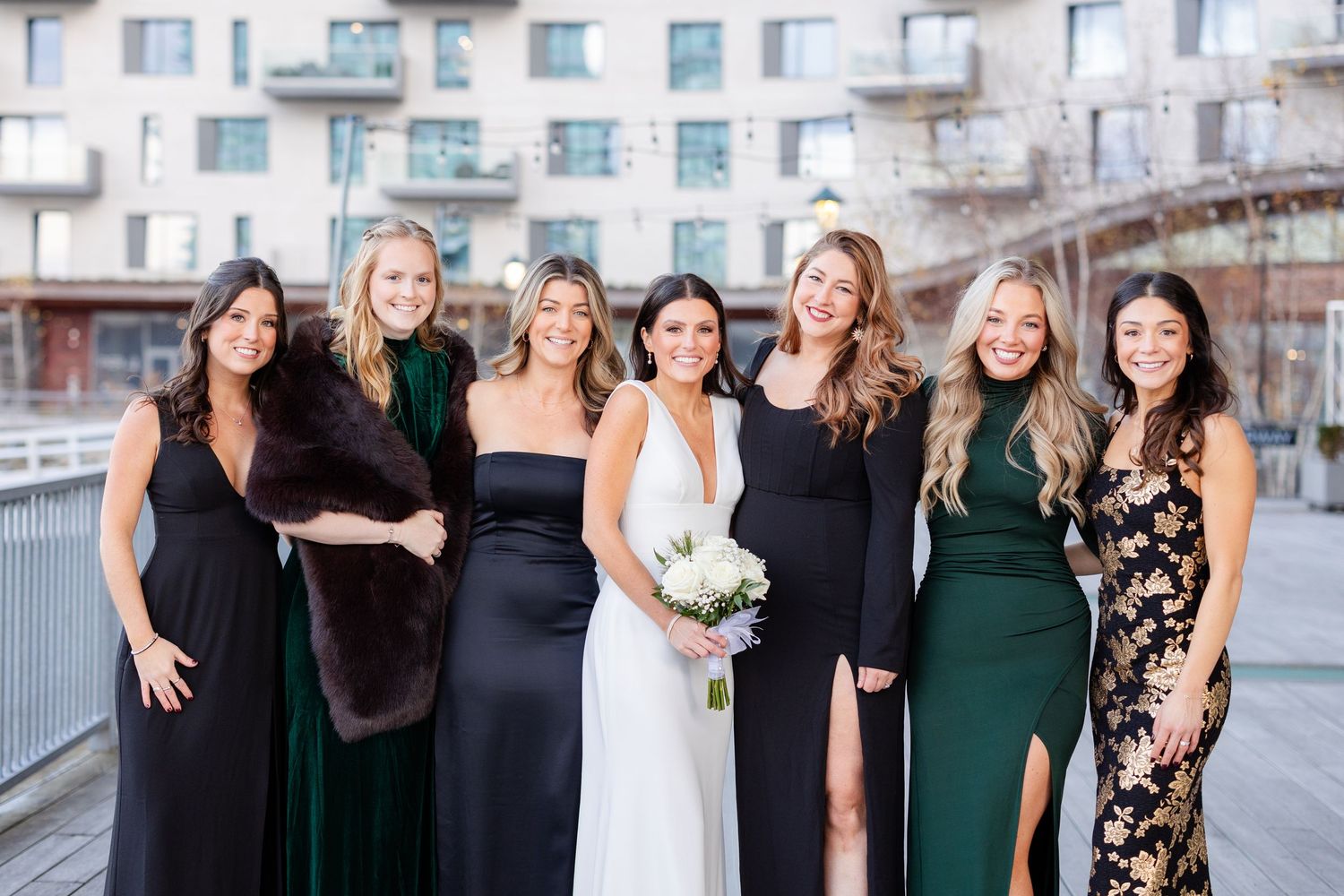 Bride with bridesmaids in black and green dresses posing outdoors at urban wedding venue.