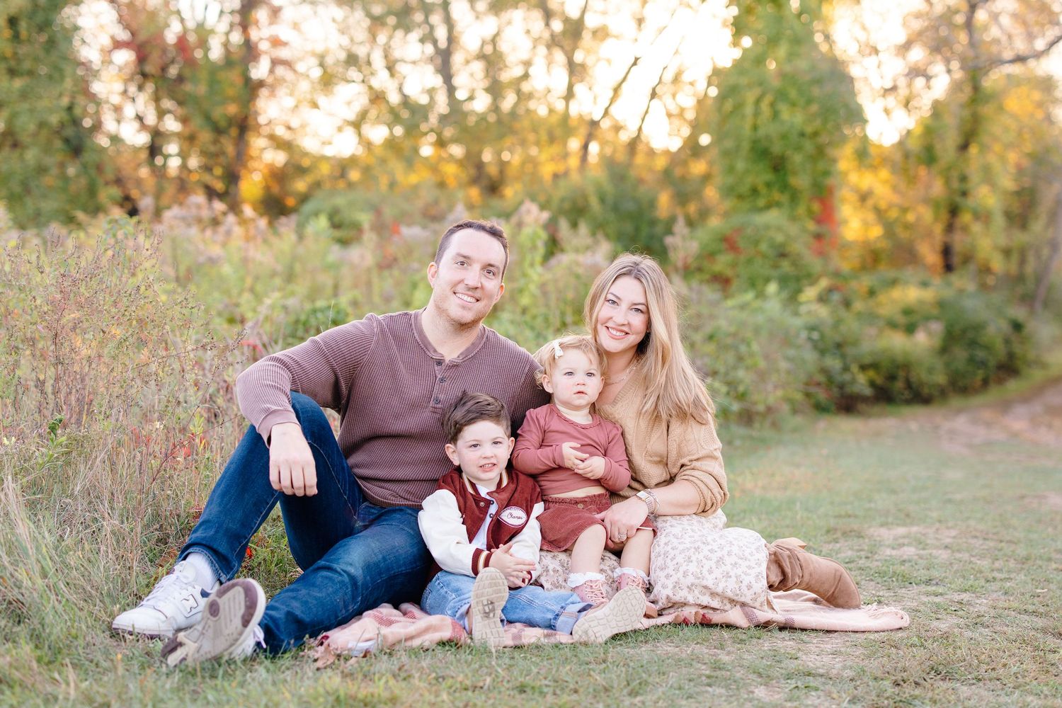 Happy family of four sitting together in a sunlit autumn meadow with colorful foliage in the background.