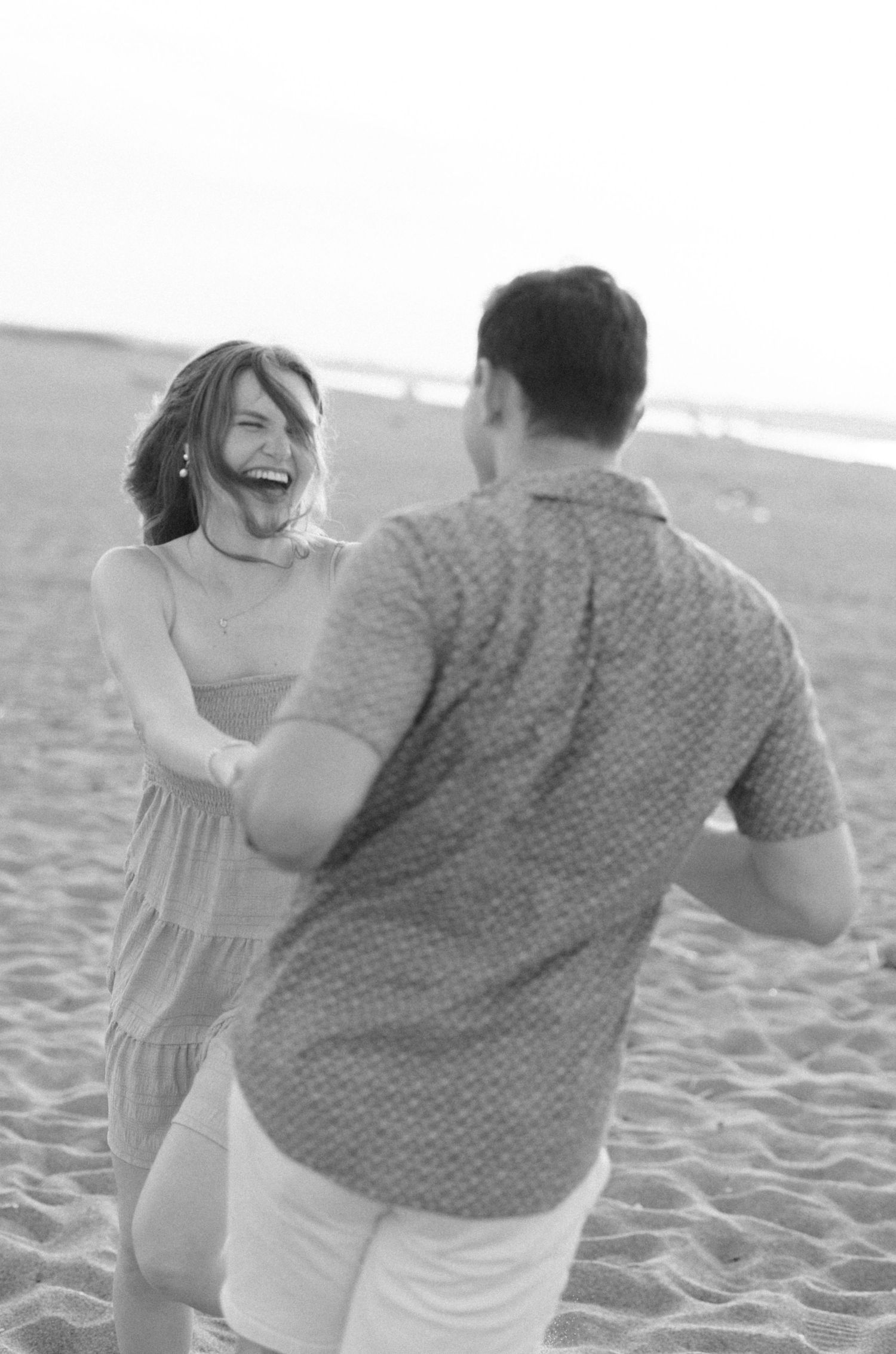 A black and white photo of a laughing woman dancing with a man on a sandy beach during an engagement session.