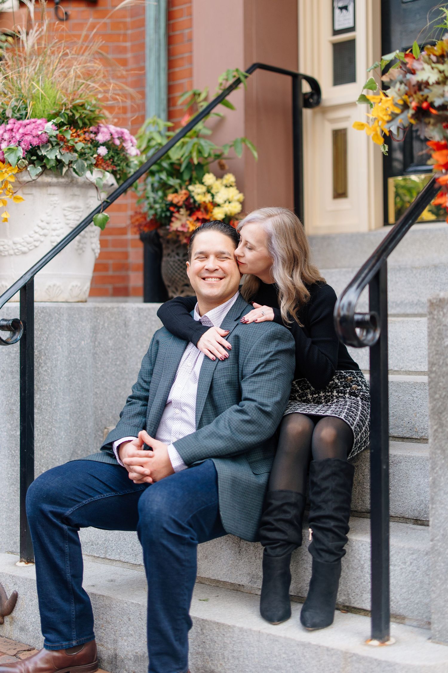 A happy couple sits on stone steps surrounded by colorful autumn flowers, the woman kissing the mans cheek.