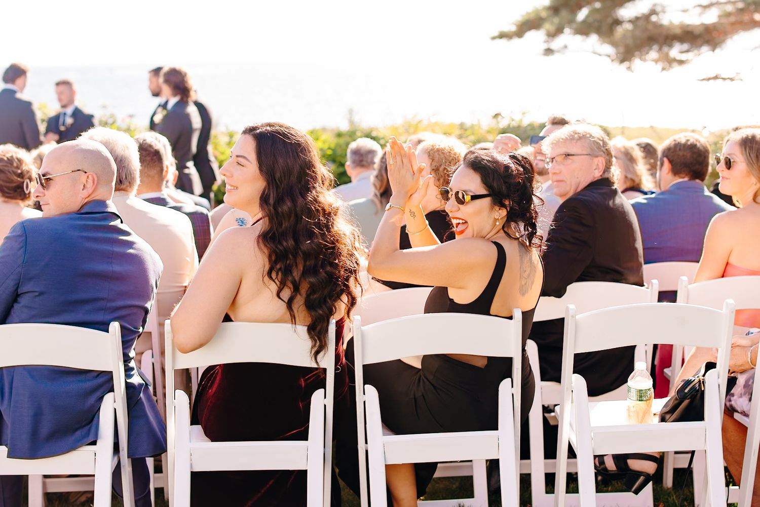 Wedding guests seated in white chairs at outdoor ceremony, two women in black dresses smiling.