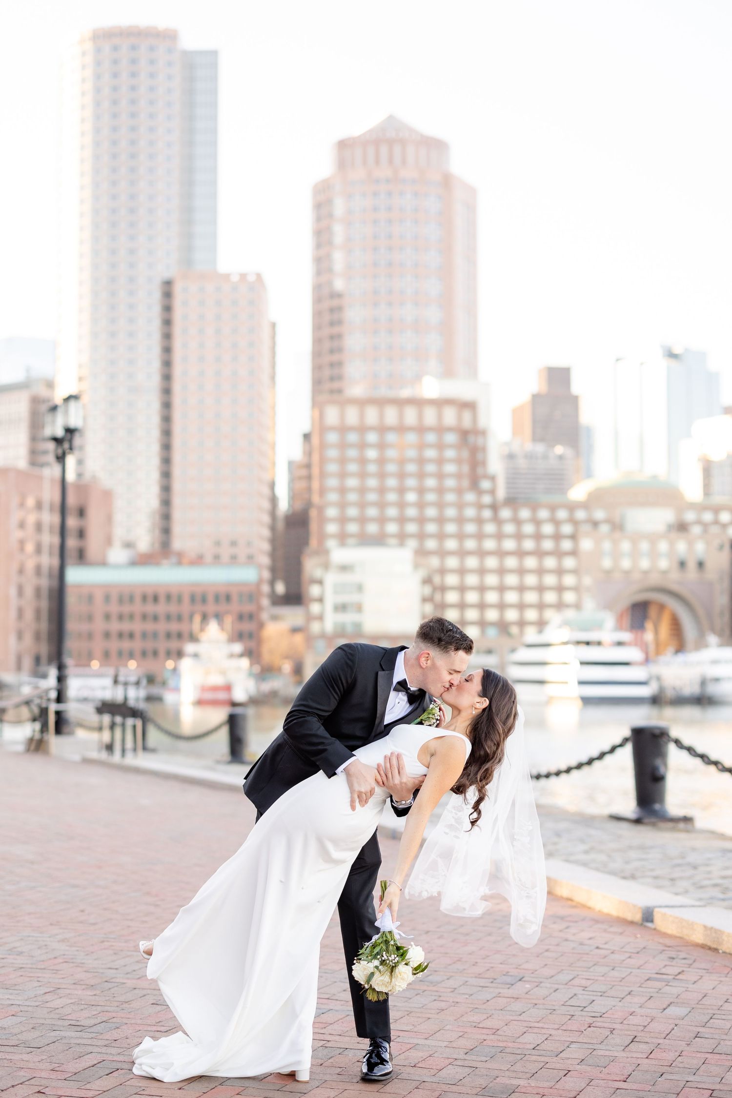 Groom dipping bride in wedding kiss on brick waterfront with Boston city skyline at golden hour.