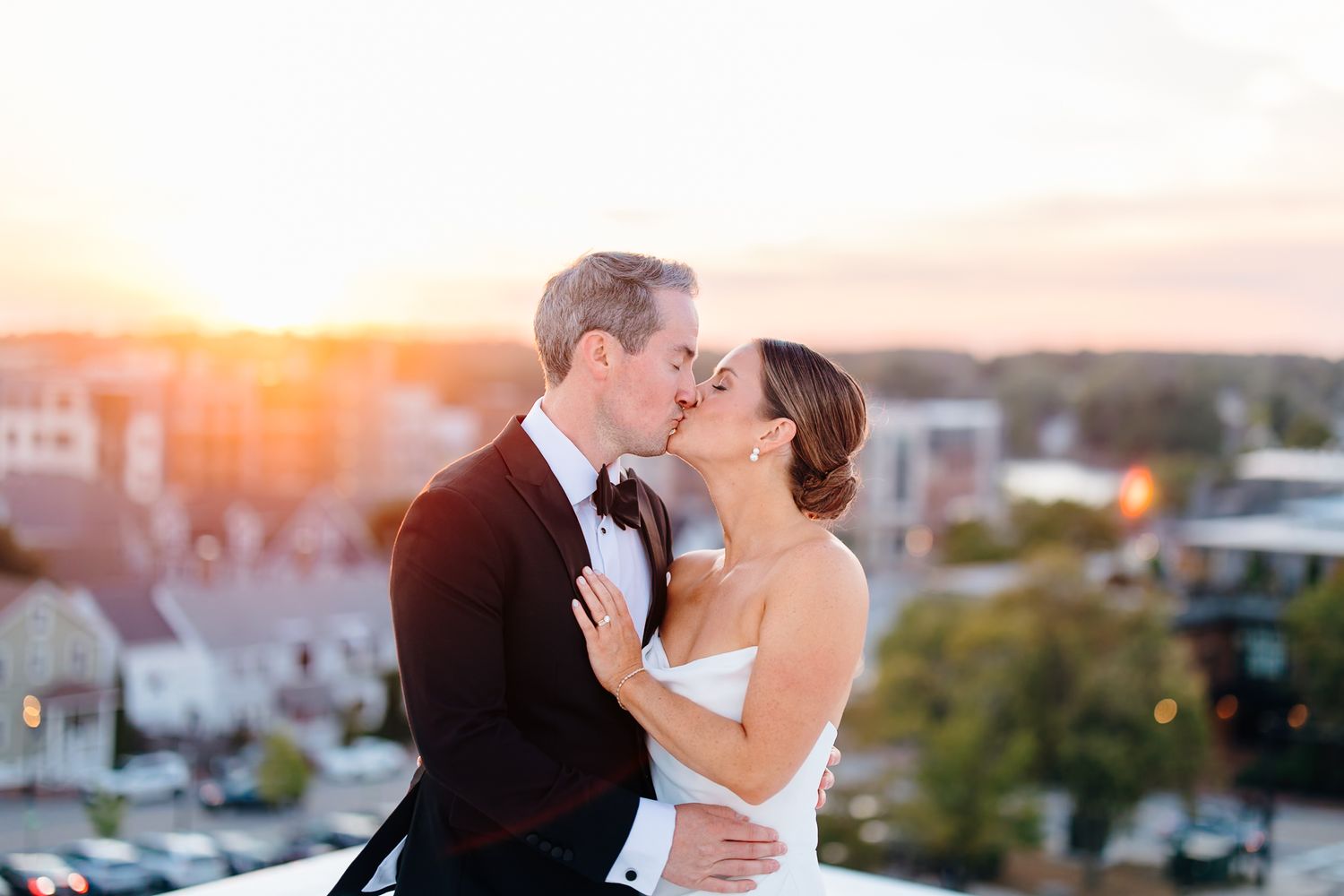 Bride and groom share a romantic rooftop kiss at sunset with a city skyline backdrop on their wedding day.