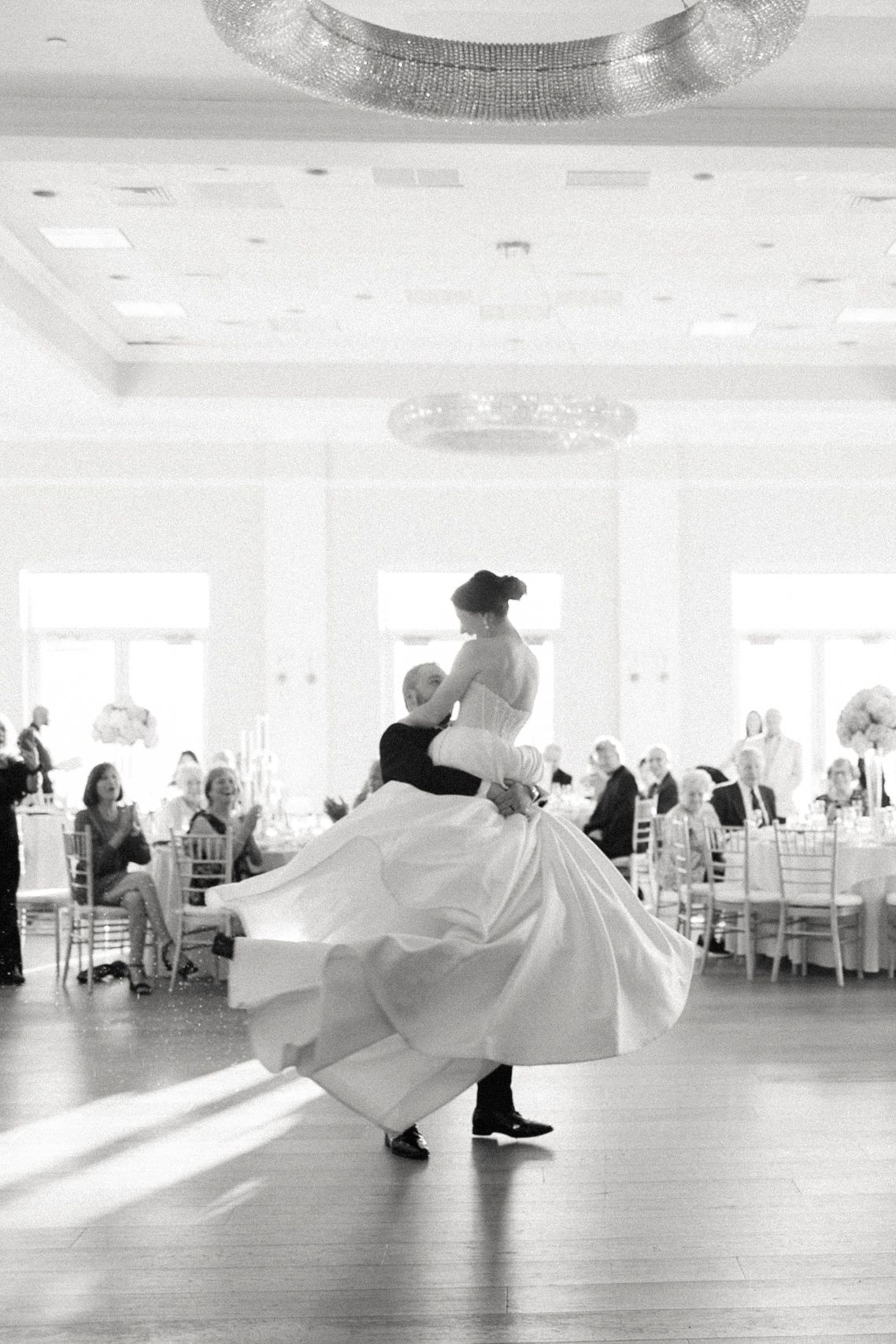 Black and white photo of groom spinning bride during first dance in elegant ballroom.