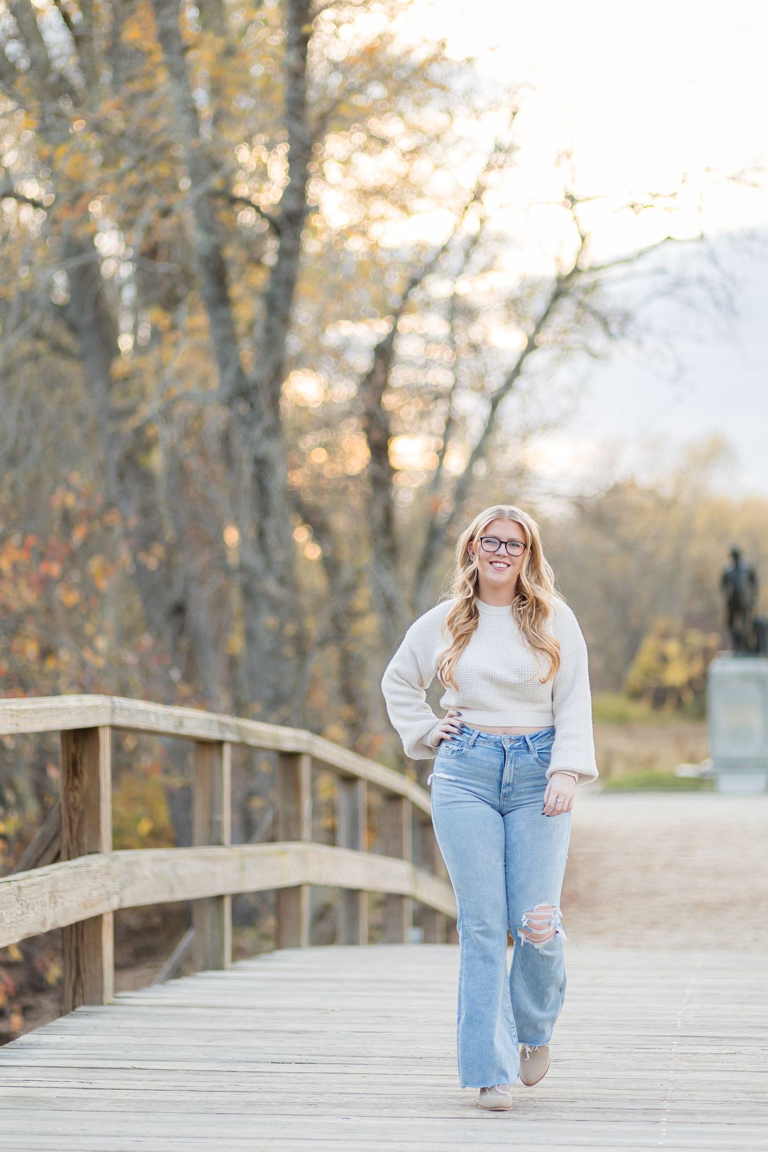 Young woman in white sweater and ripped jeans walking on wooden bridge surrounded by autumn foliage.