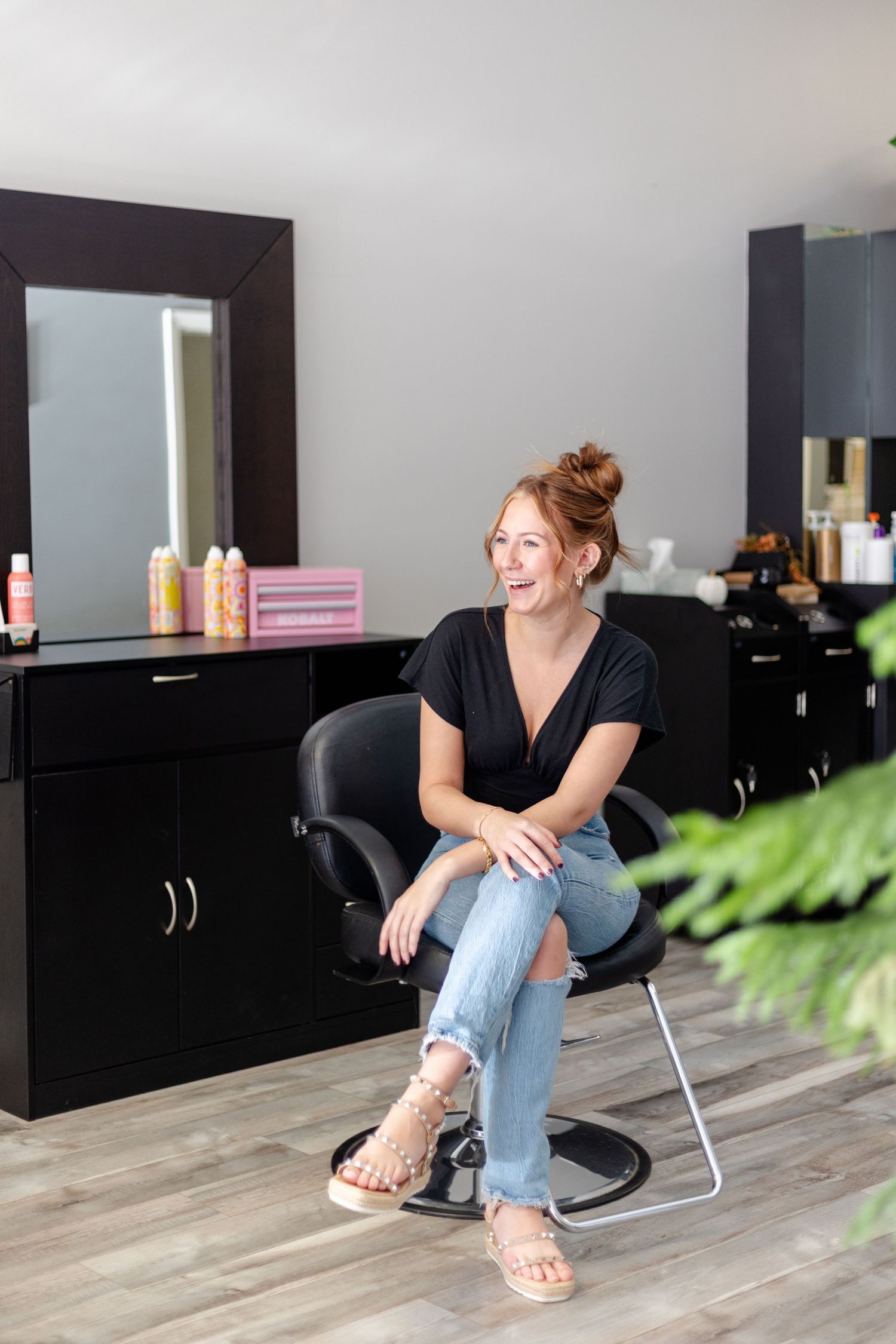 Redhead hairstylist laughing while sitting in salon chair surrounded by styling products.