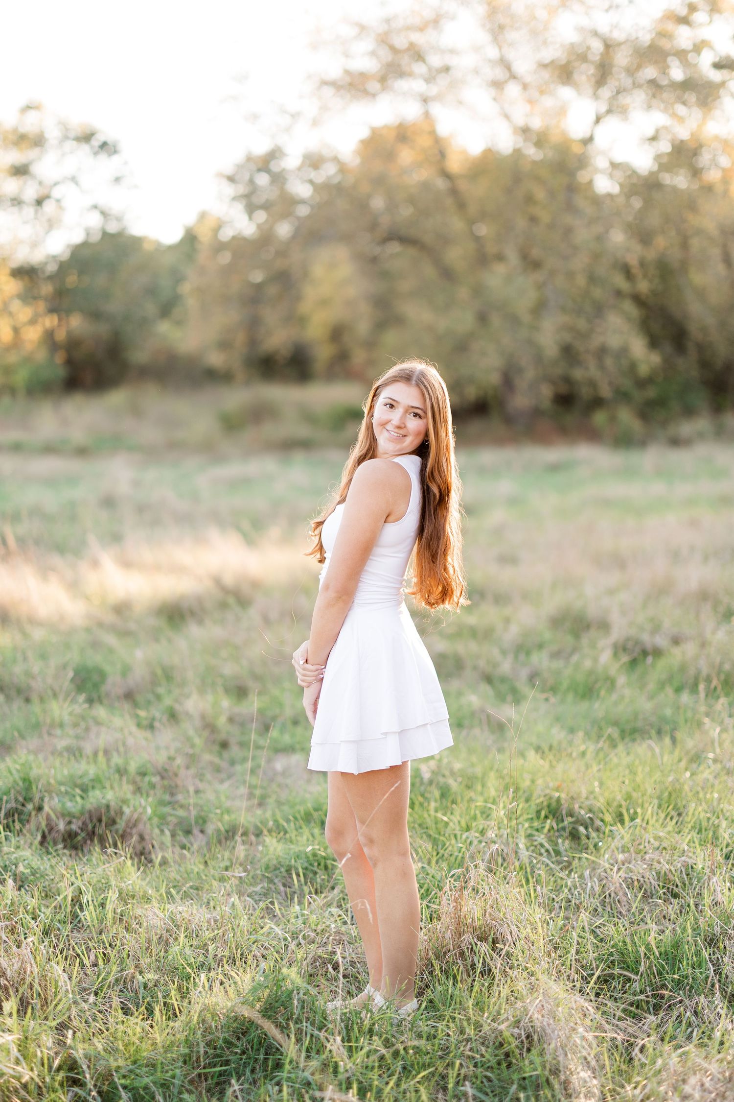 Young woman with long red hair in white mini dress standing in a sunlit field with trees in the background.