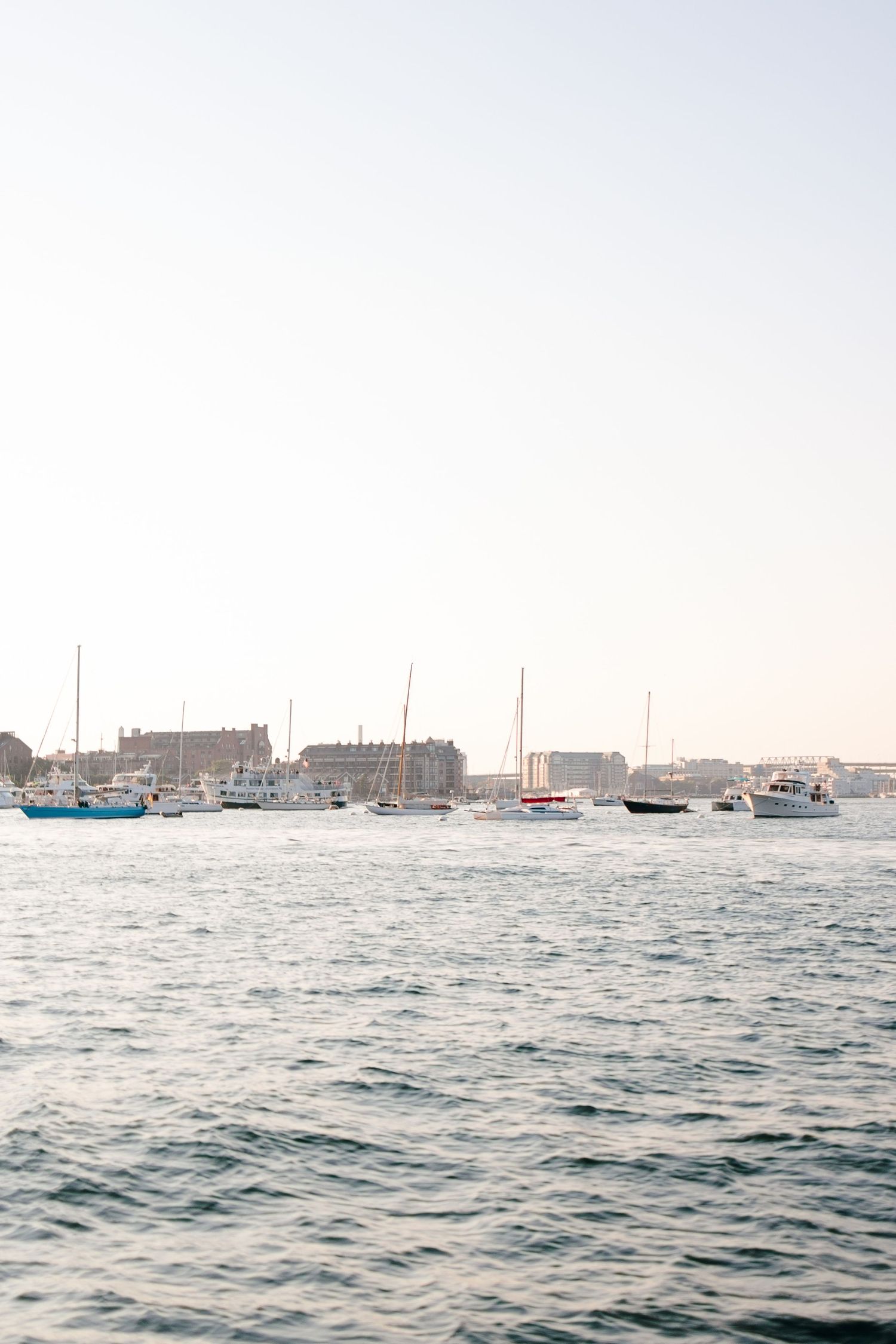Sailboats and yachts anchored in calm harbor waters at dusk with city buildings in the background.