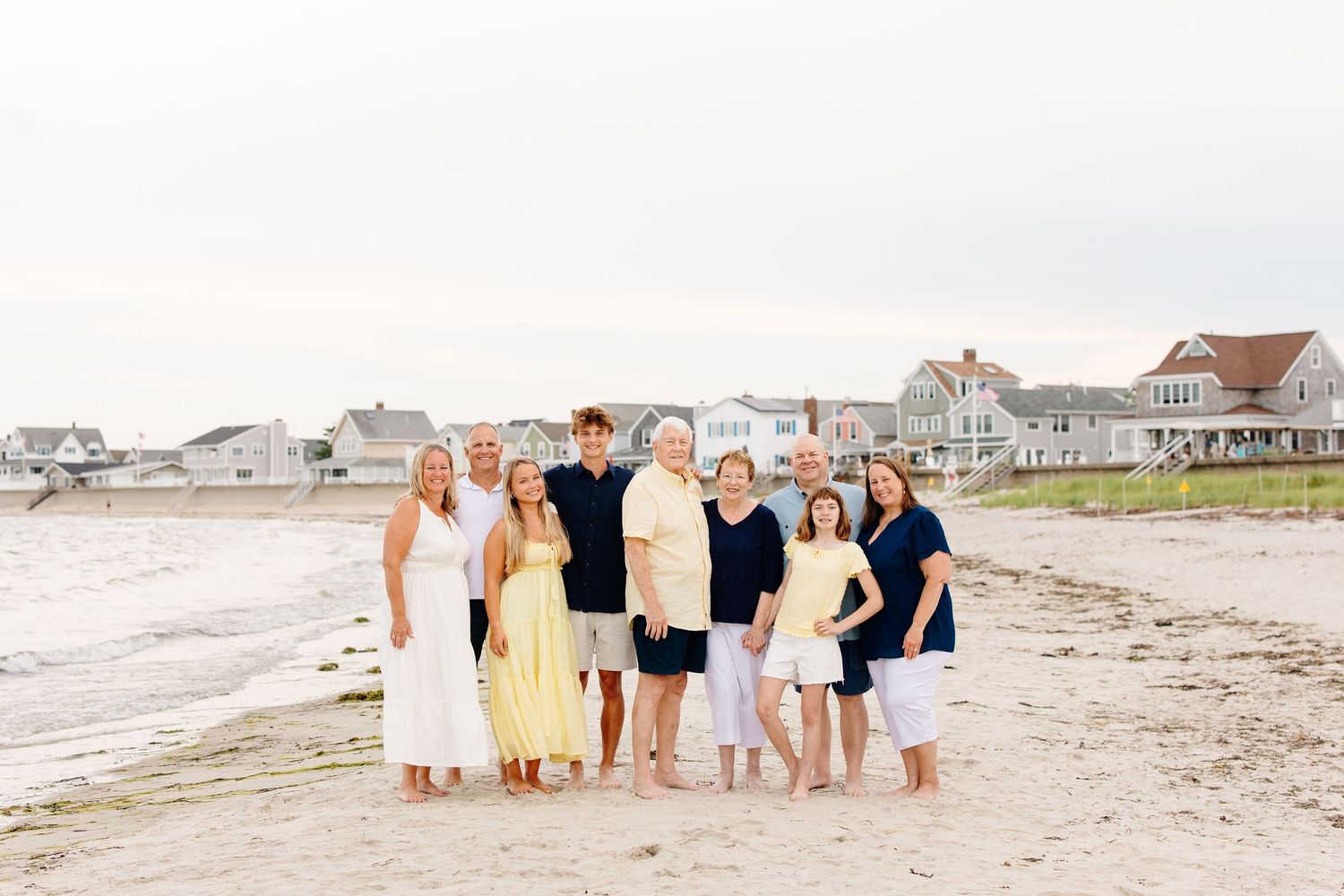 Extended family of eight poses together on a sandy beach with coastal homes visible in the background.