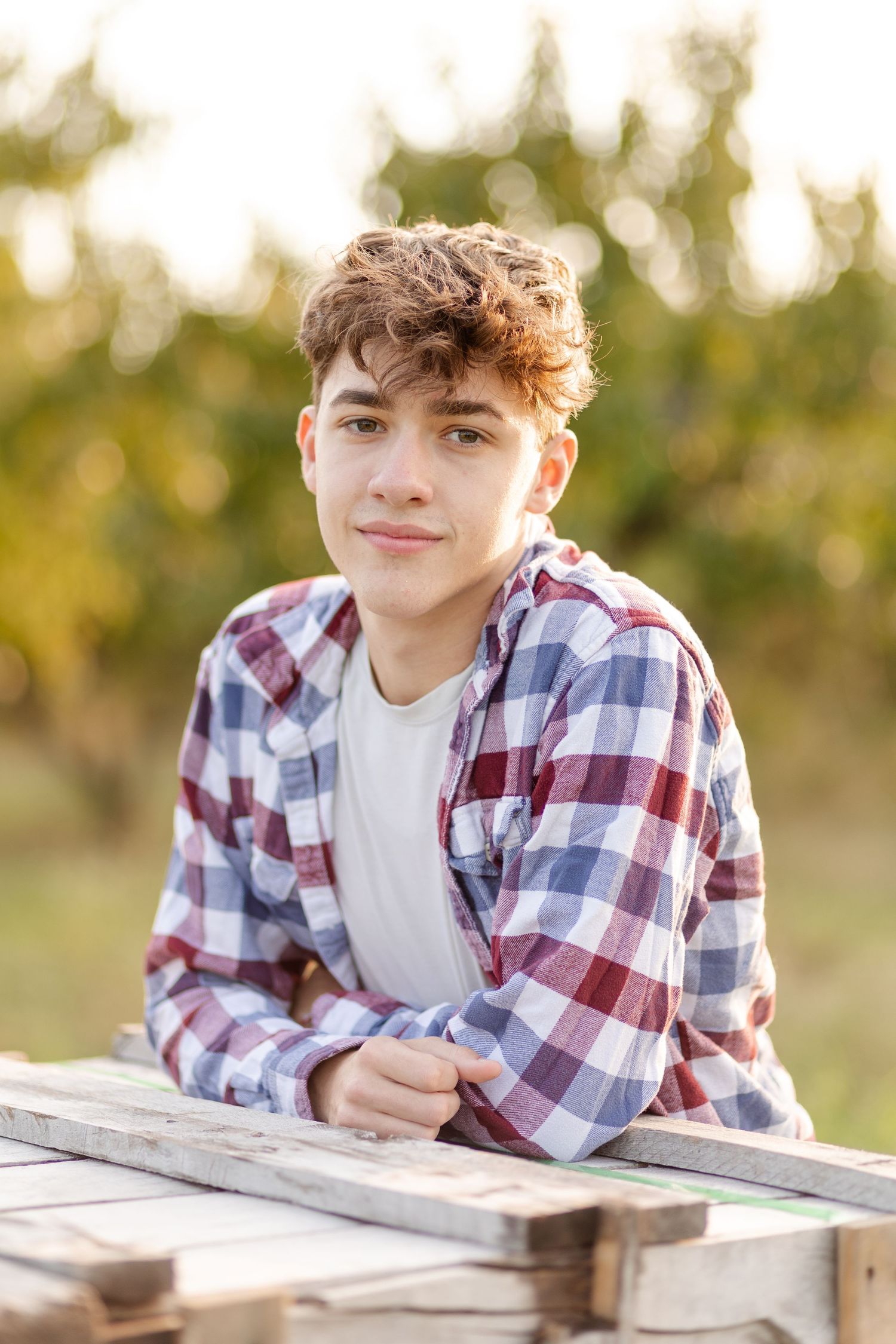 A teenage boy in a plaid shirt leaning on a wooden surface outdoors with a bokeh green background.