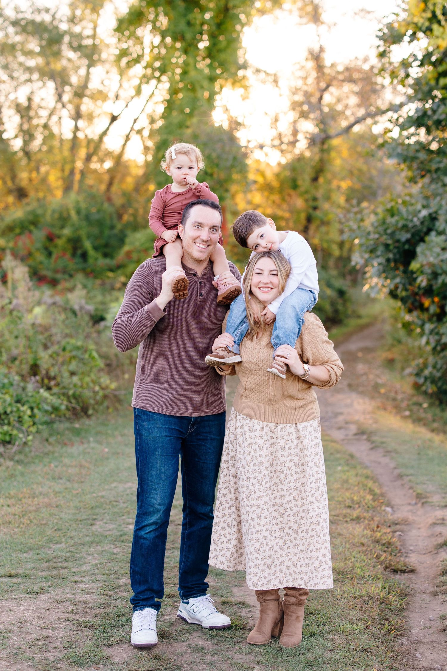 Smiling family of four with children on shoulders poses on autumn trail at golden hour.