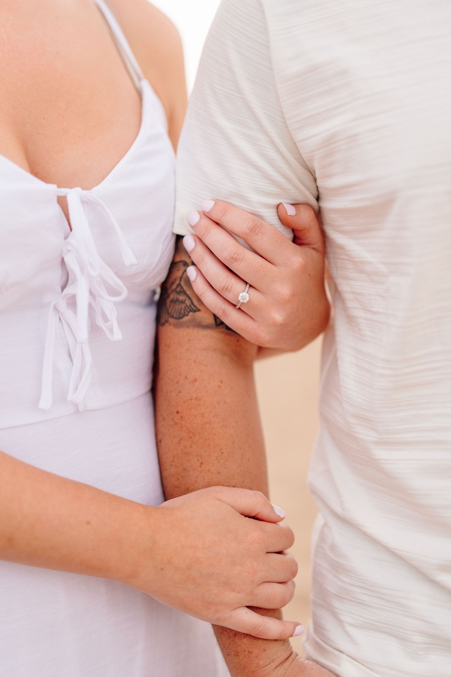 Close-up of a woman holding a mans arm on a beach, showcasing her diamond engagement ring.