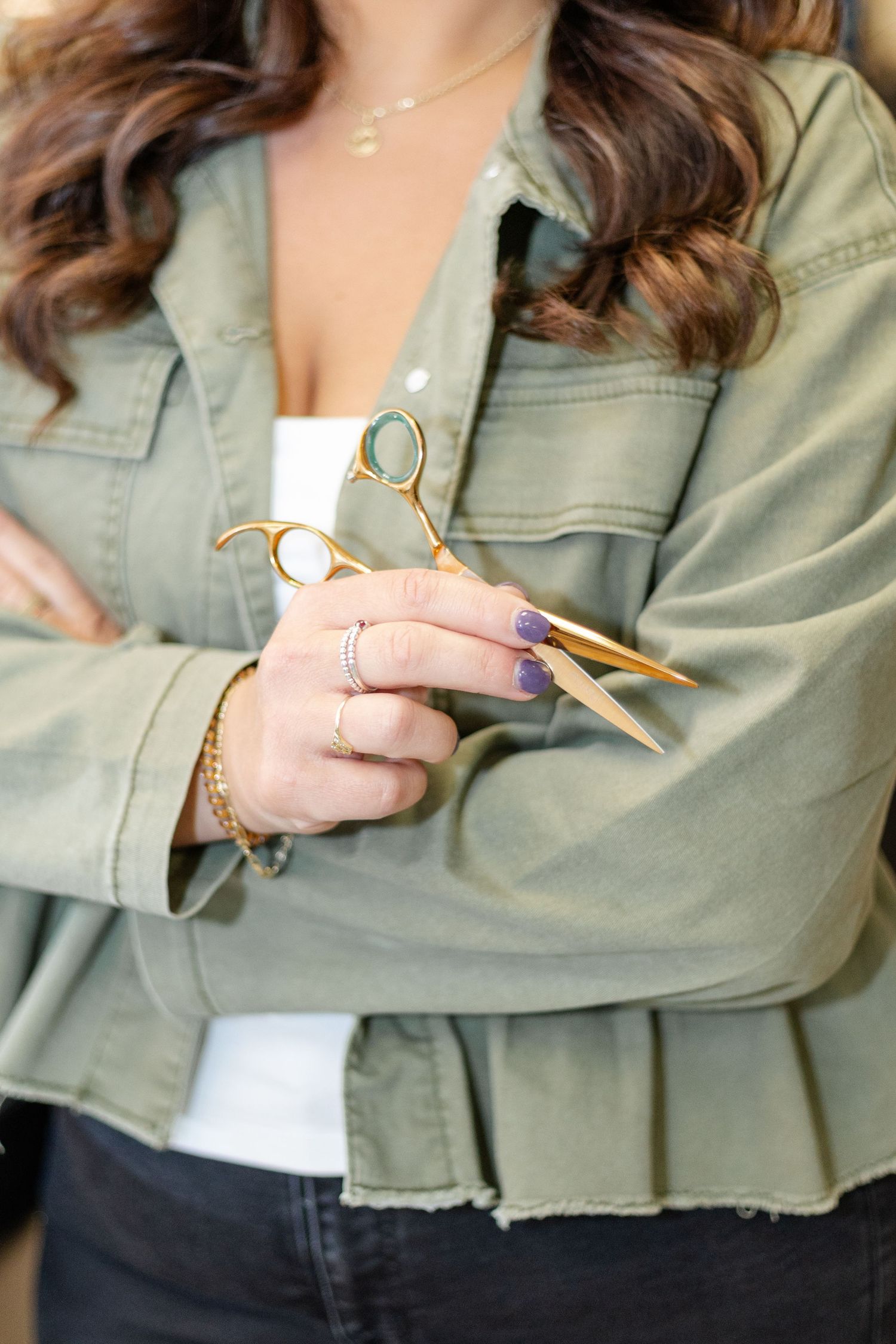 Female hairstylist in olive jacket holding gold scissors with purple manicured nails.