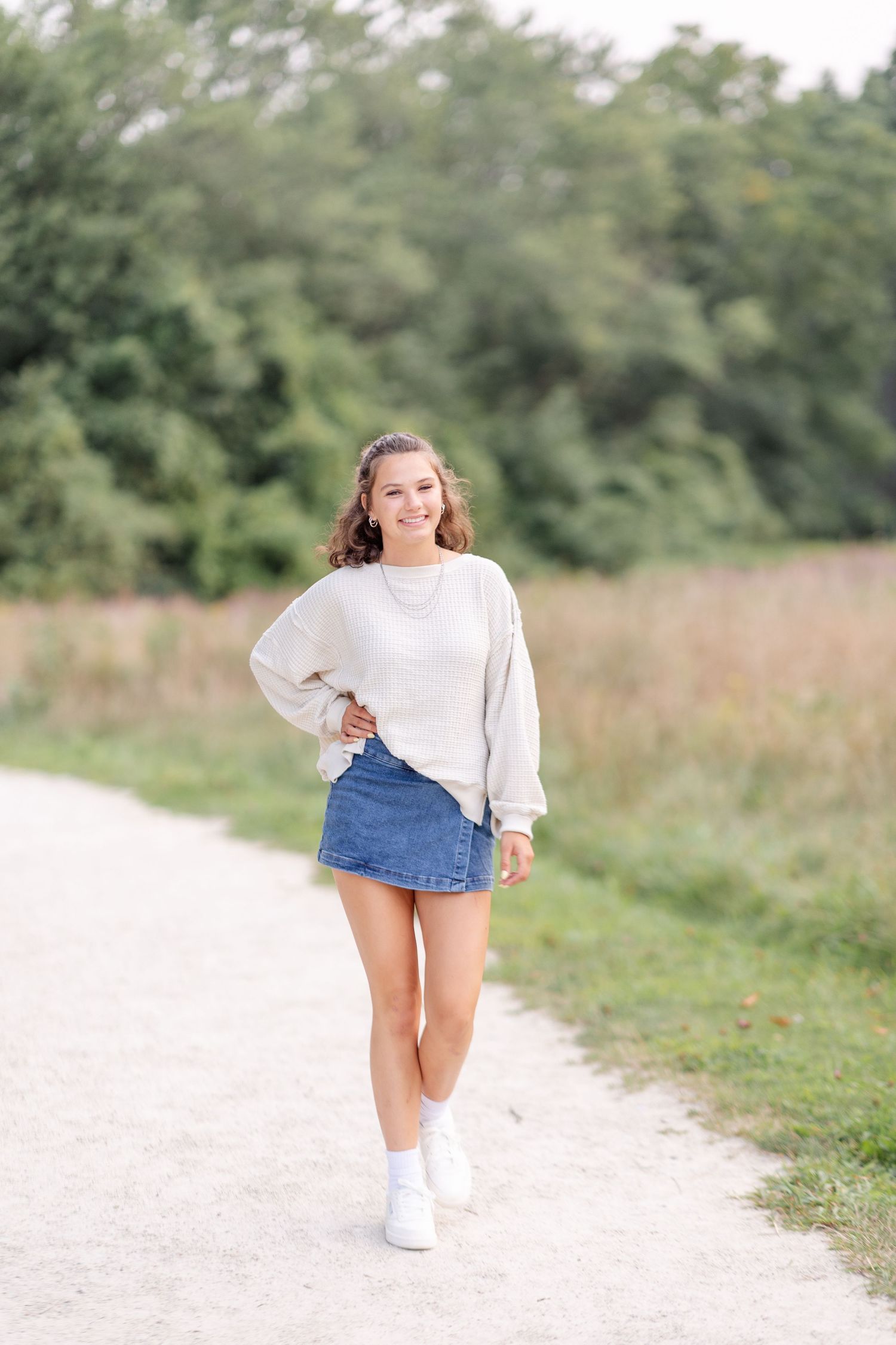 Smiling young woman in gray sweatshirt and denim skirt walking on gravel path near greenery.
