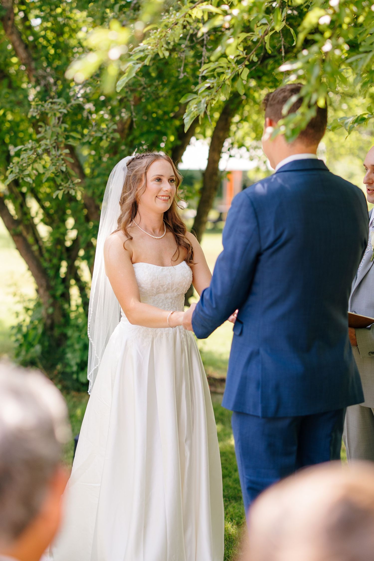 Bride in white lace gown and veil smiles while holding hands with groom during outdoor ceremony.