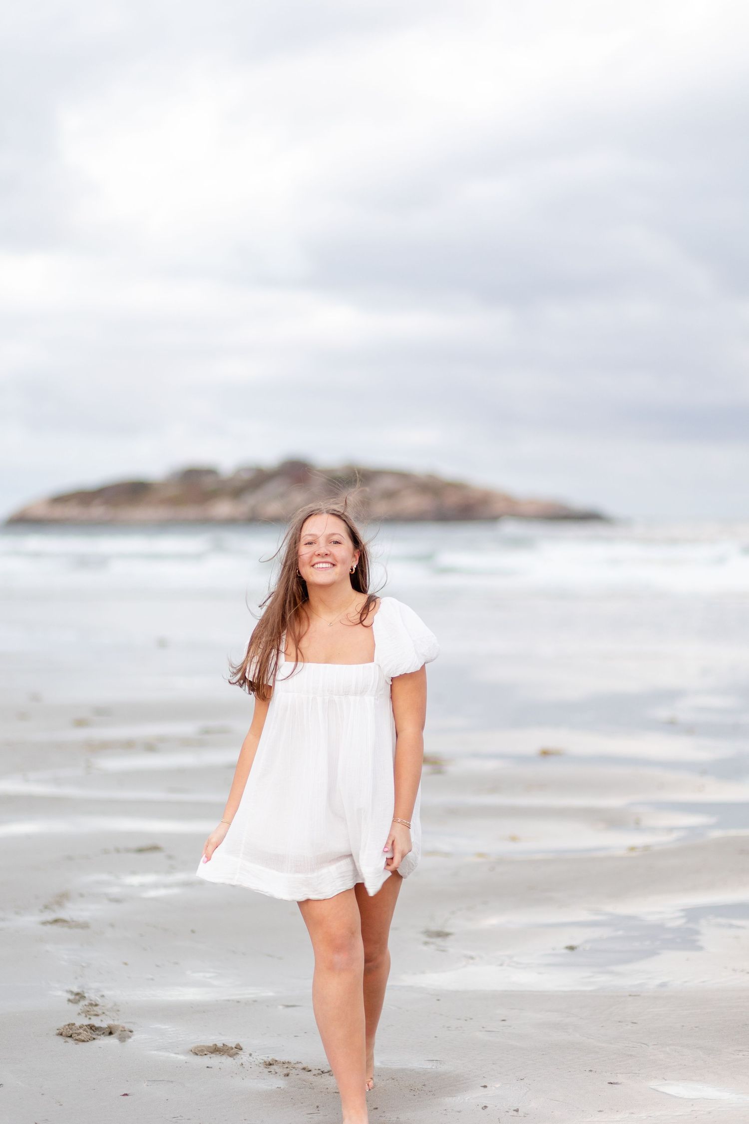 Smiling young woman in white dress walking along ocean shoreline on a cloudy day.
