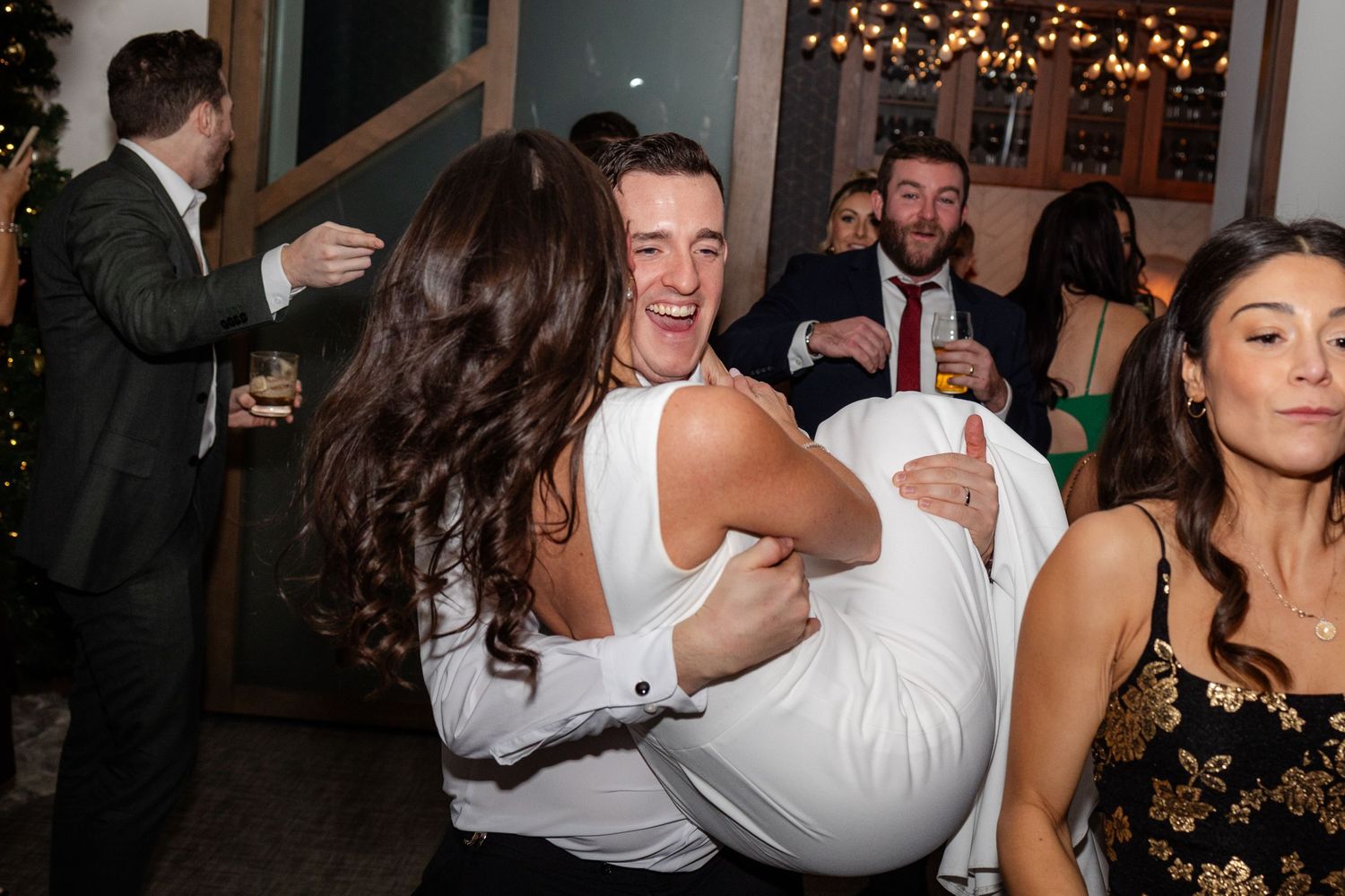 Groom joyfully lifts bride during lively wedding reception dance floor celebration with guests in background.