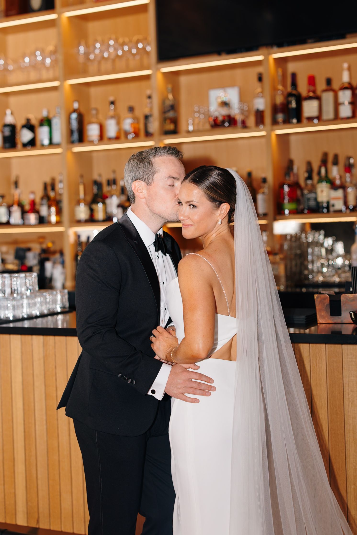 Groom kissing bride on cheek as she smiles at camera in front of warmly lit bar backdrop.