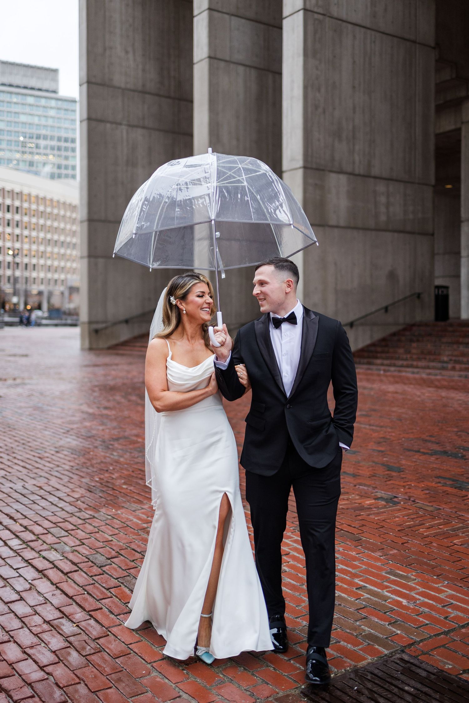 Bride and groom walking under clear bubble umbrella on rainy brick street in Boston.