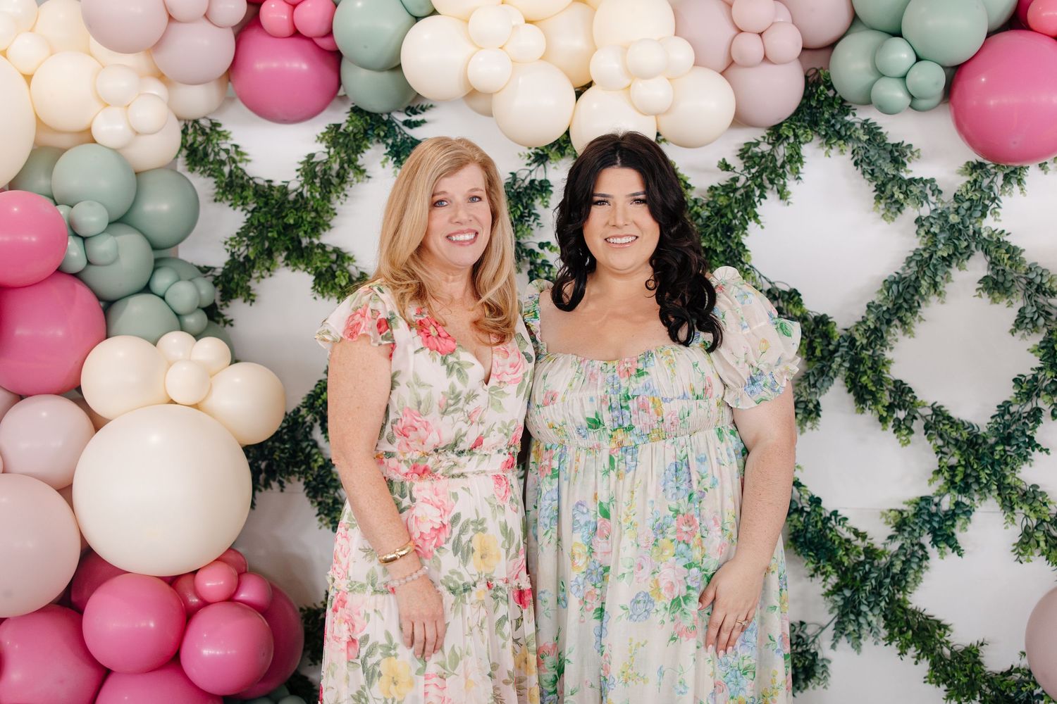 Two women in floral dresses smiling before colorful balloon and greenery backdrop.