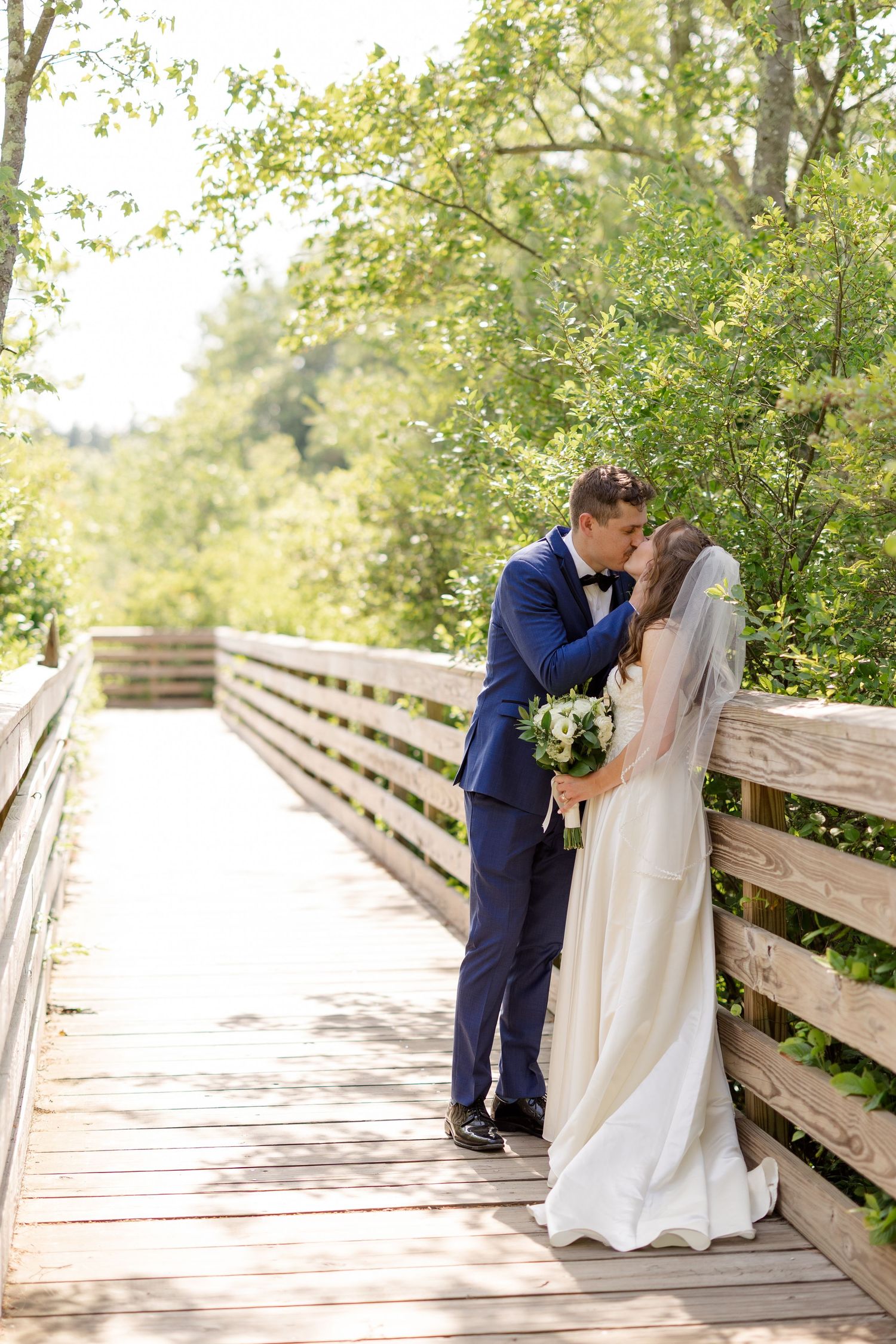Bride and groom share a romantic kiss on a wooden bridge surrounded by lush green trees on their wedding day.