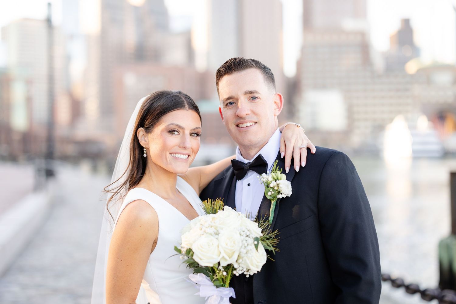 A bride holding white flowers and a groom in a tuxedo smiling together with a city skyline in the background.