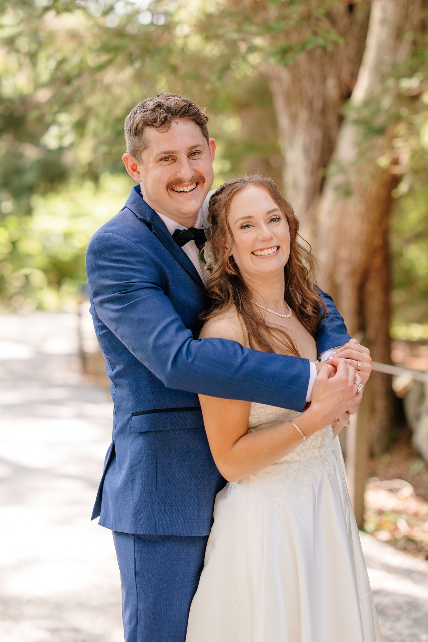 Smiling groom in blue suit embraces bride in white gown outdoors among trees on their wedding day.
