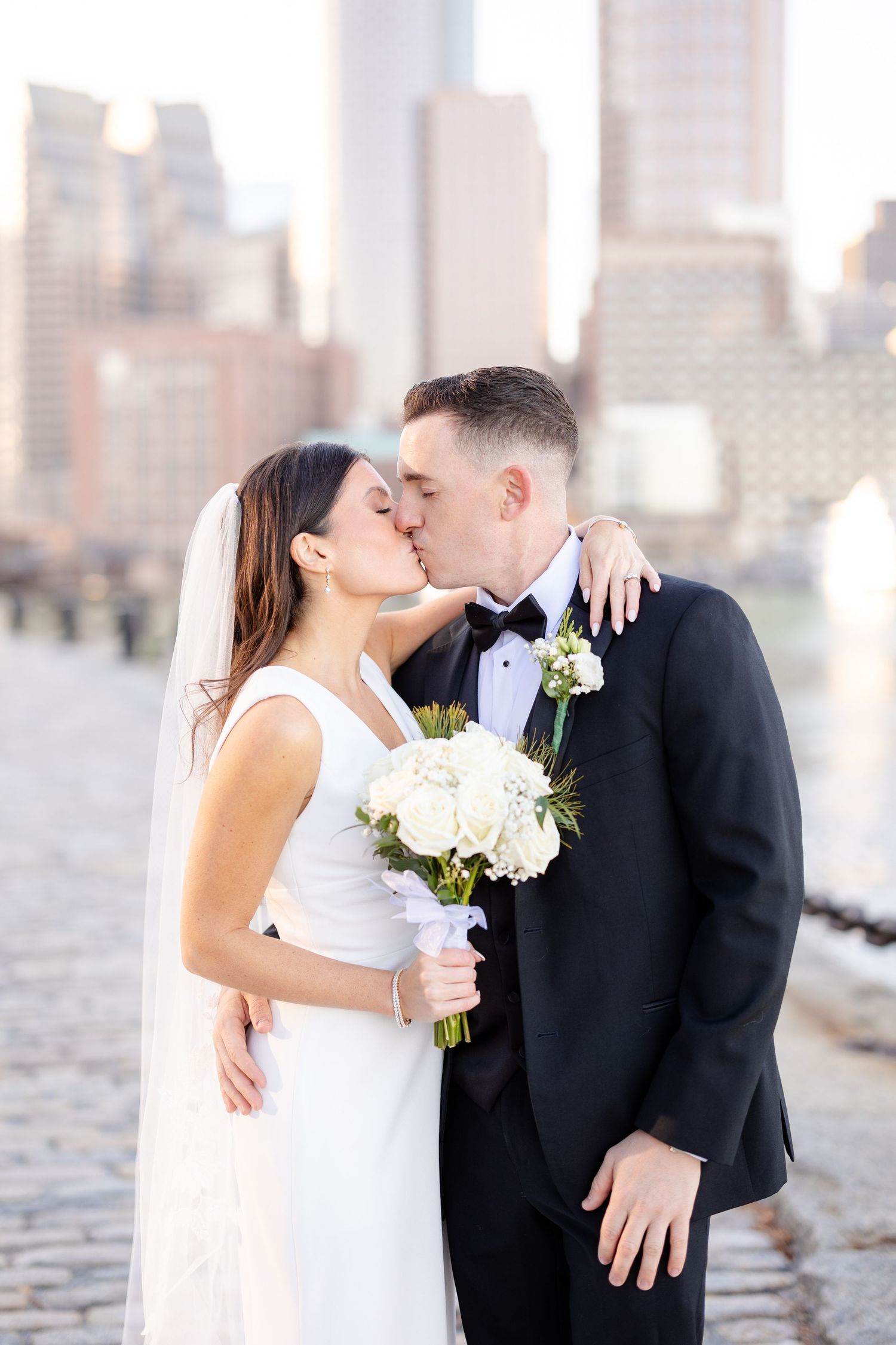 Bride and groom kiss while holding white floral bouquet with Boston waterfront skyline in background.