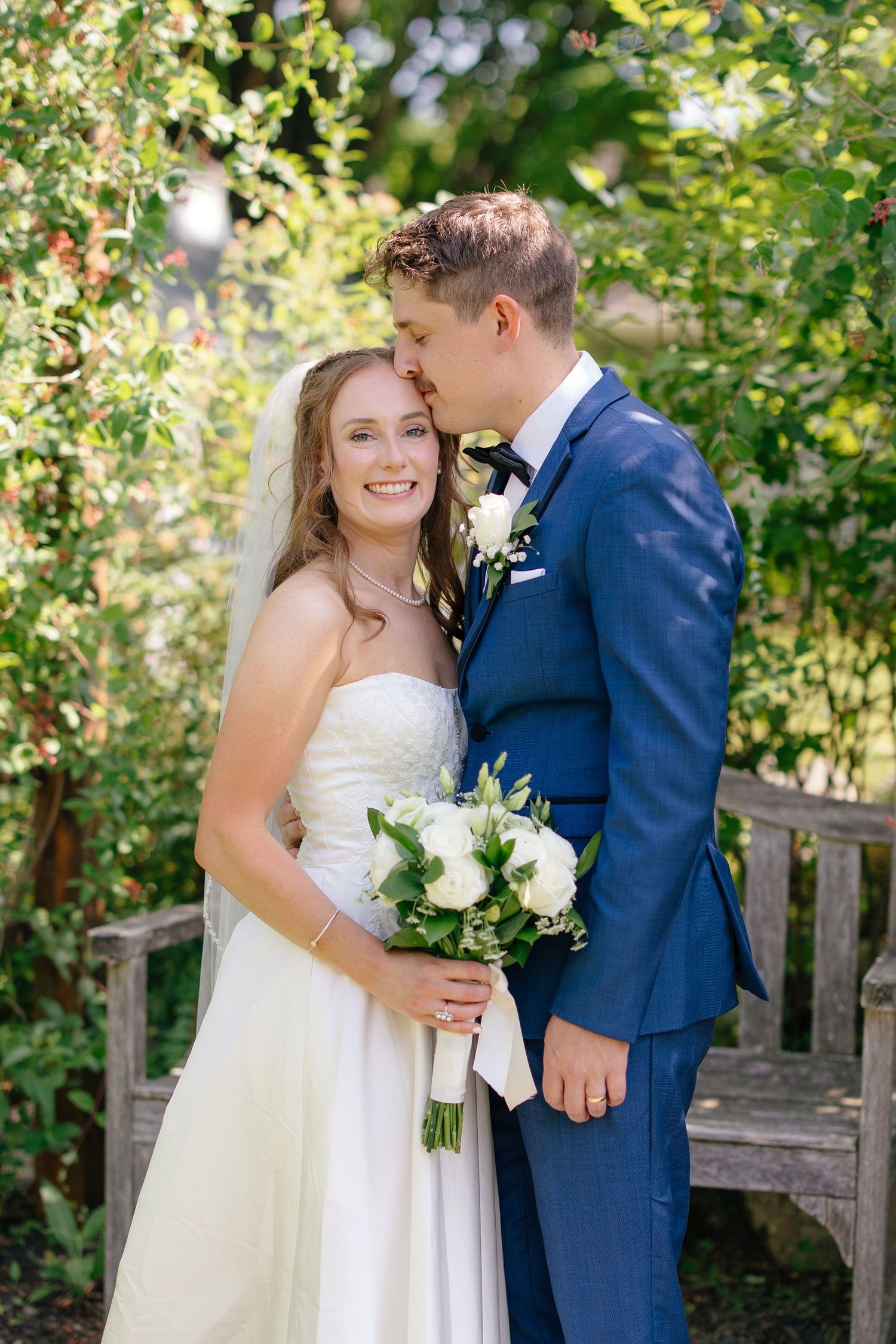 Groom in blue suit kissing bride on the cheek as she smiles holding white roses in a garden.