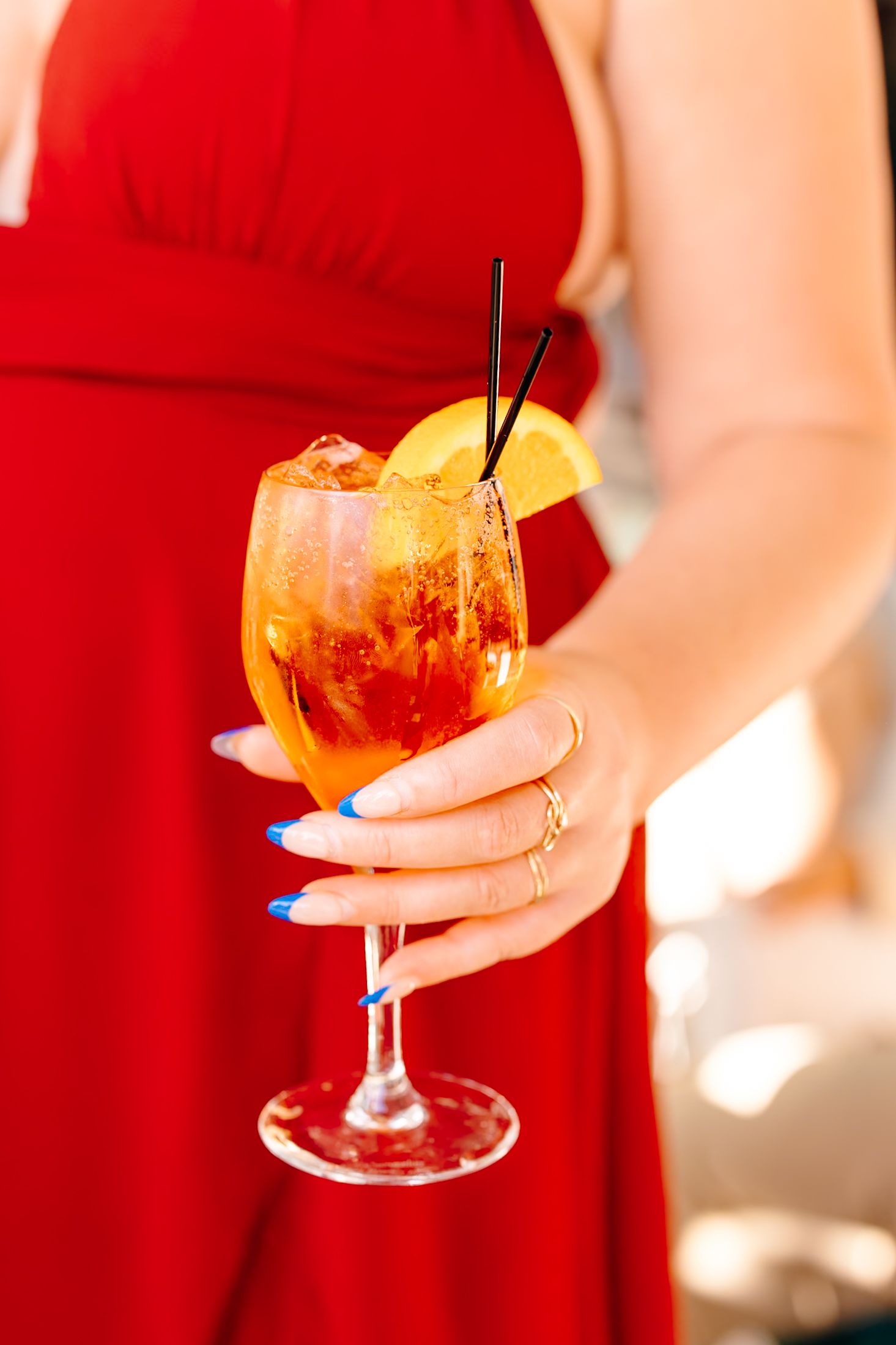 Woman in red dress holding an Aperol Spritz cocktail with orange slice, ice, and straws in a wine glass.