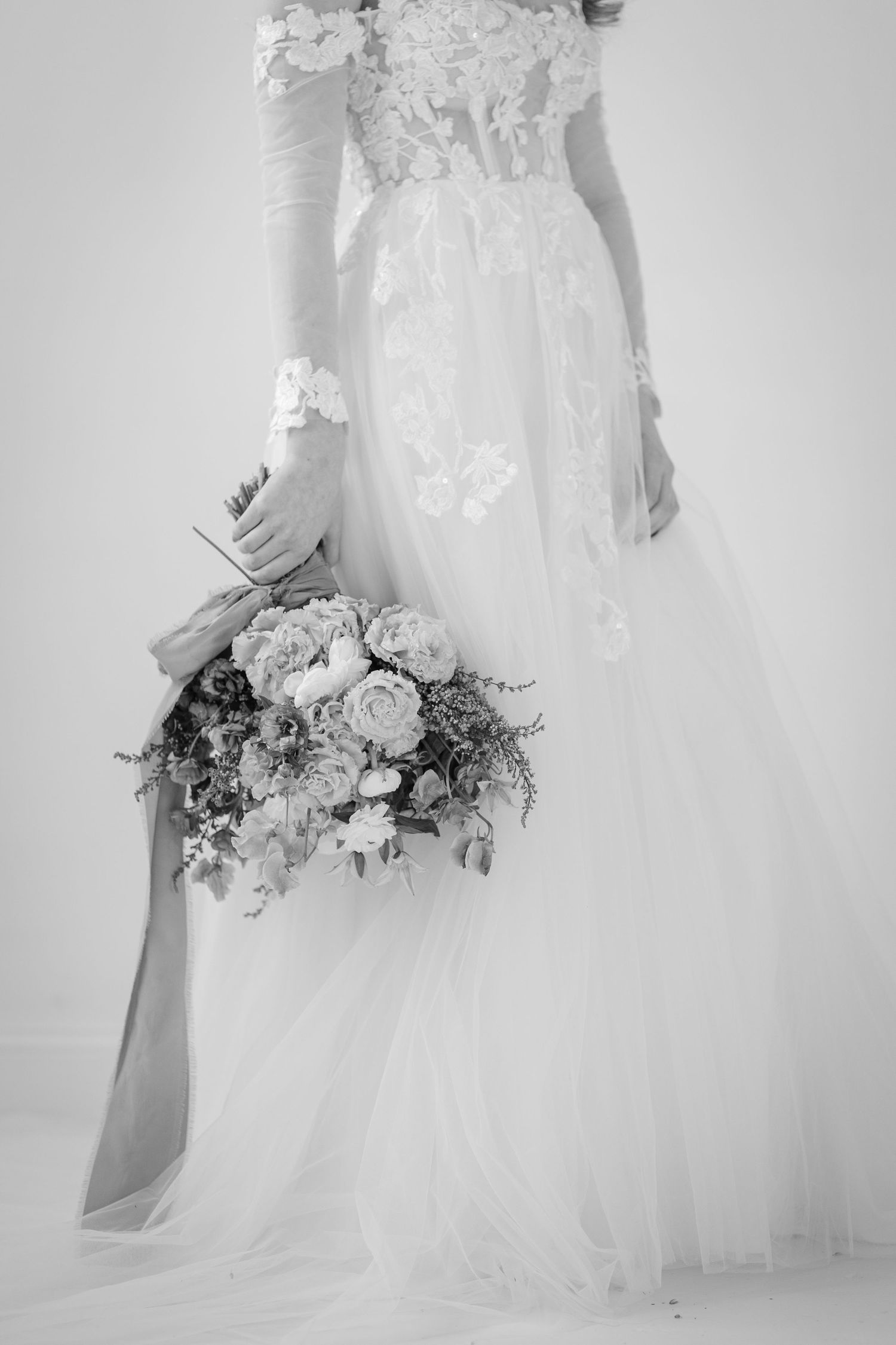 Black and white photo of bride in lace tulle gown holding elegant floral bouquet with ribbon.
