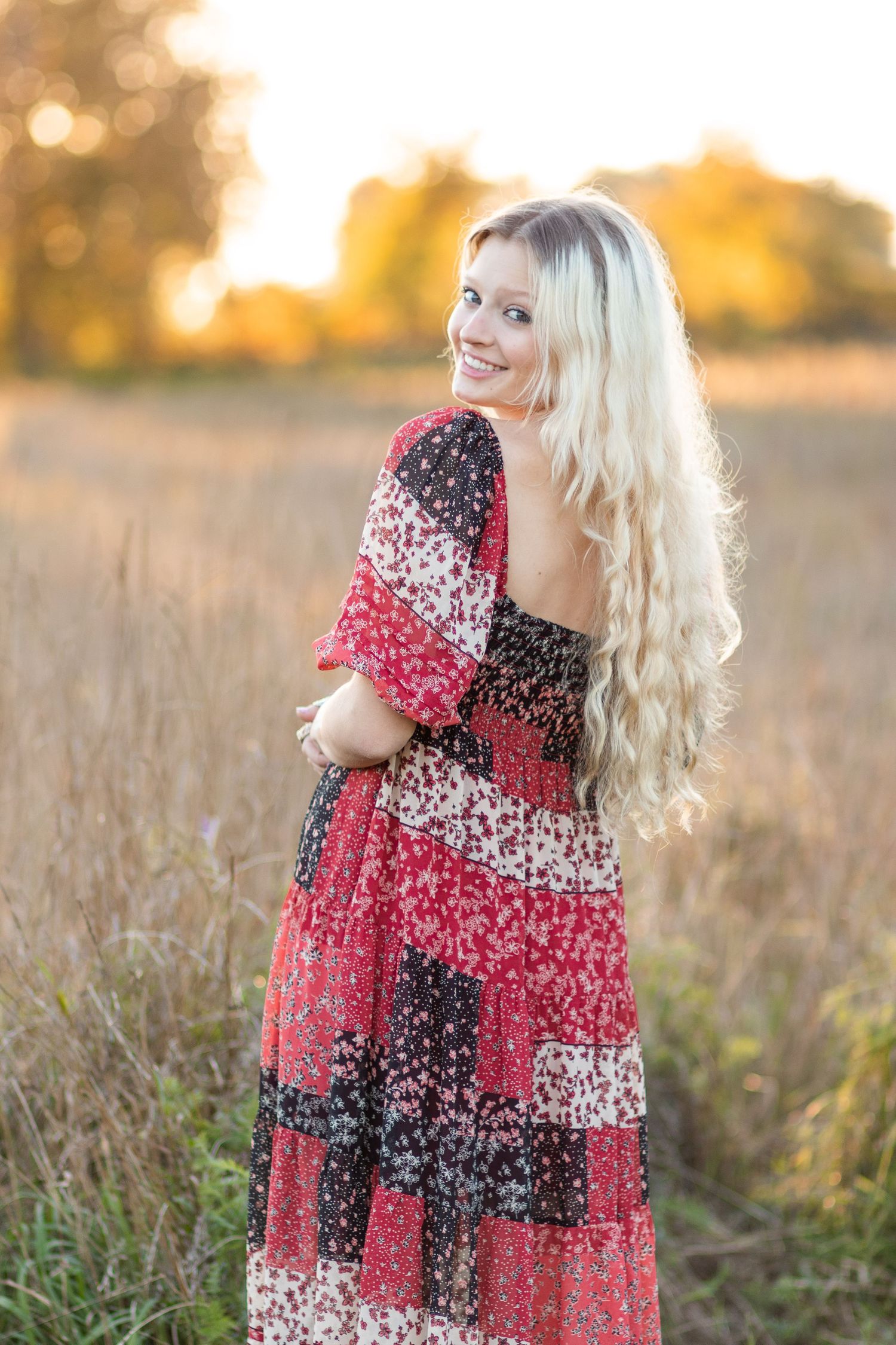 Smiling blonde woman in red patchwork maxi dress standing in a golden field at sunset.