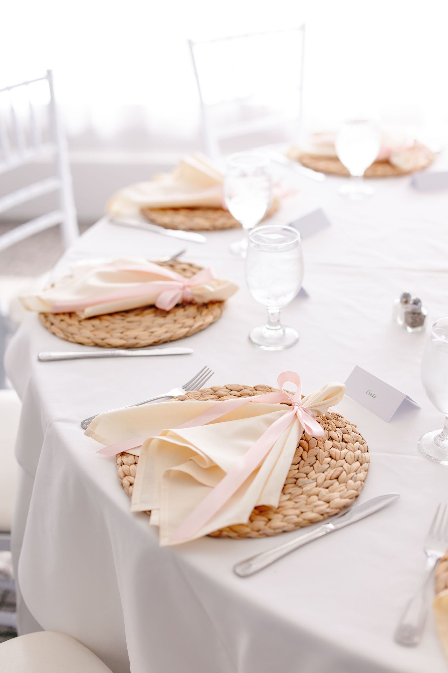 Elegant wedding table setting with woven placemats, cream napkins tied with pink ribbon, and crystal glassware.
