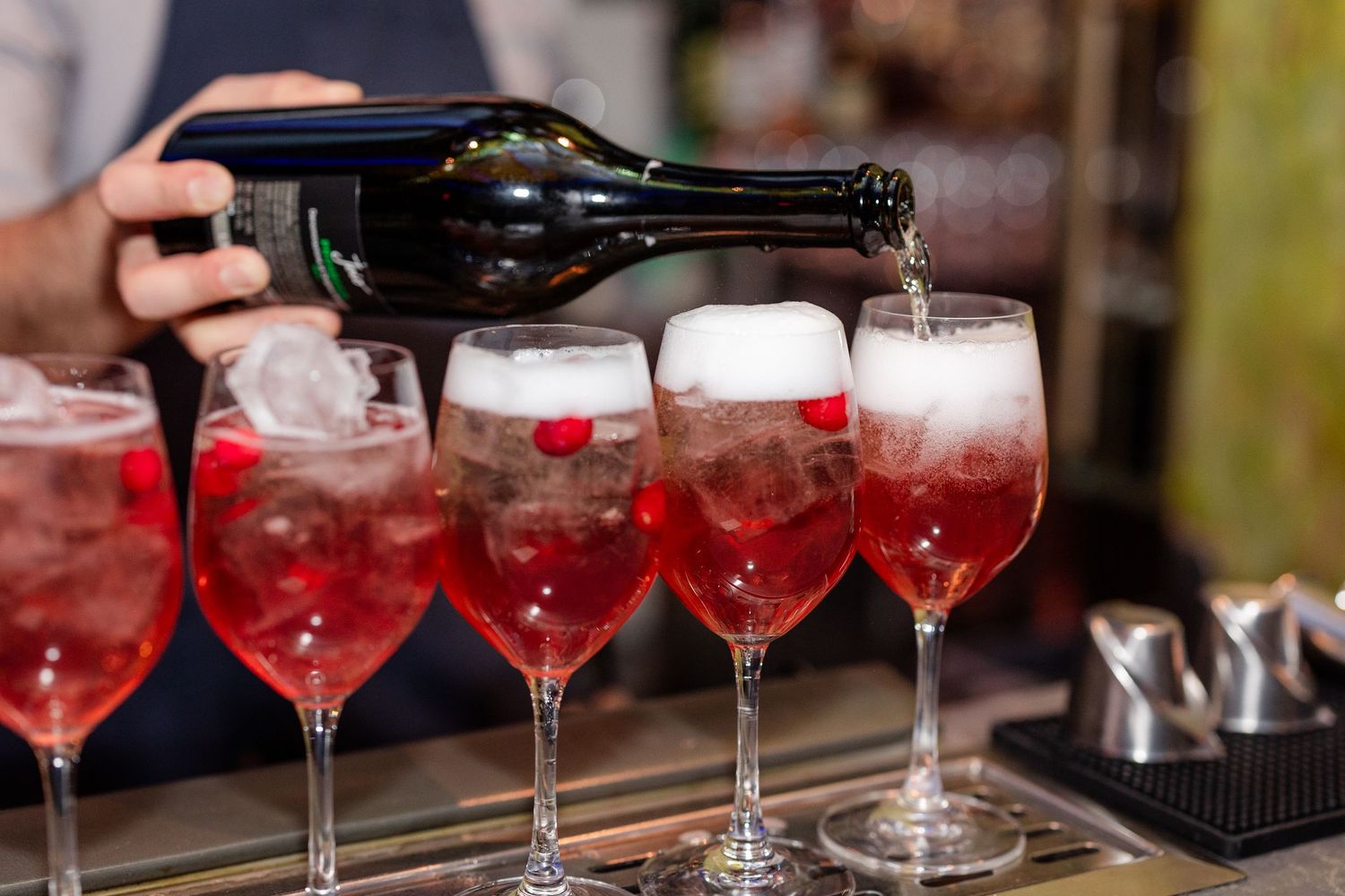 Bartender pouring champagne into red cocktail filled wine glasses at wedding bar.