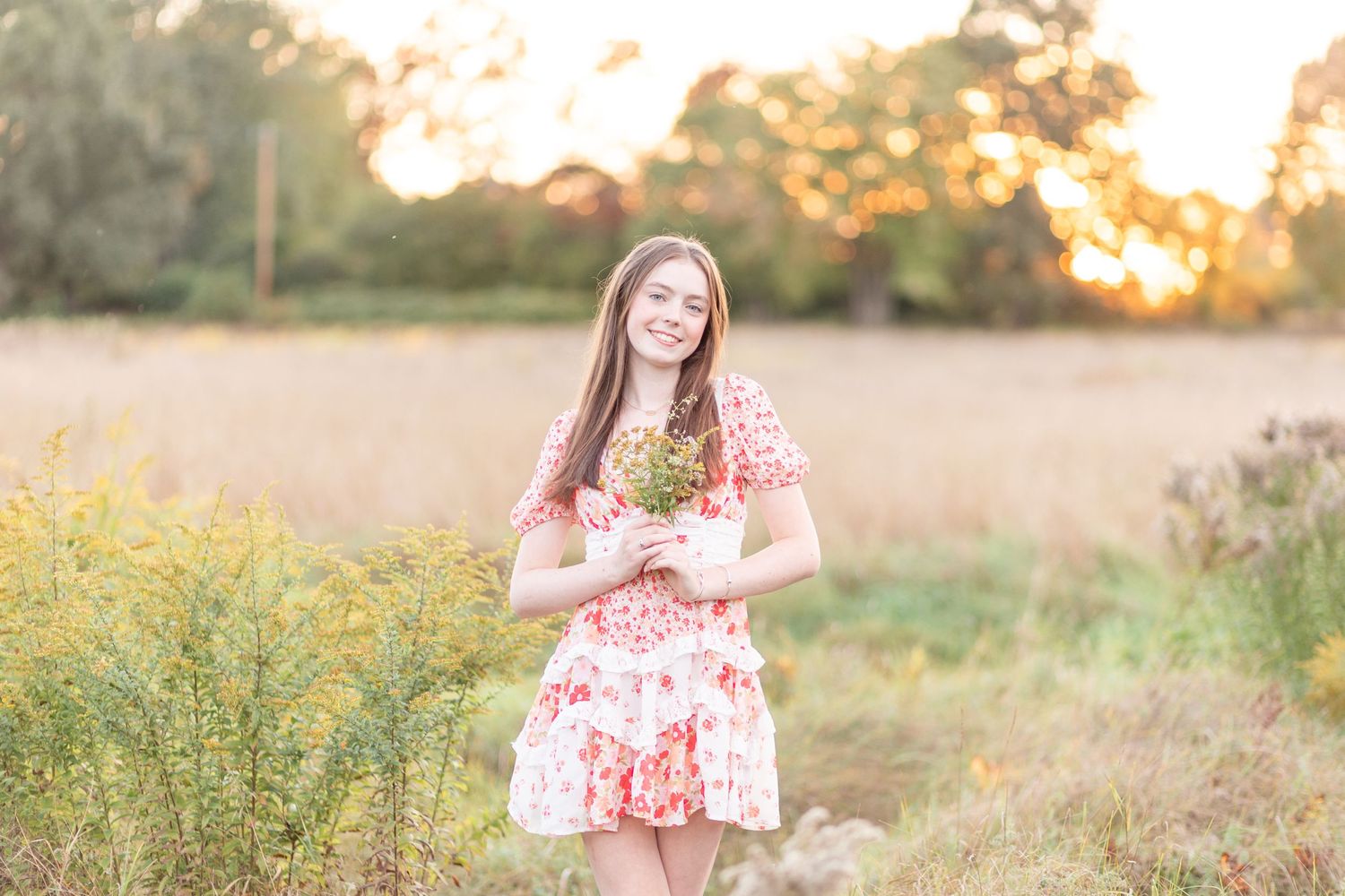 Young woman in floral dress holding wildflowers in a golden field at sunset.
