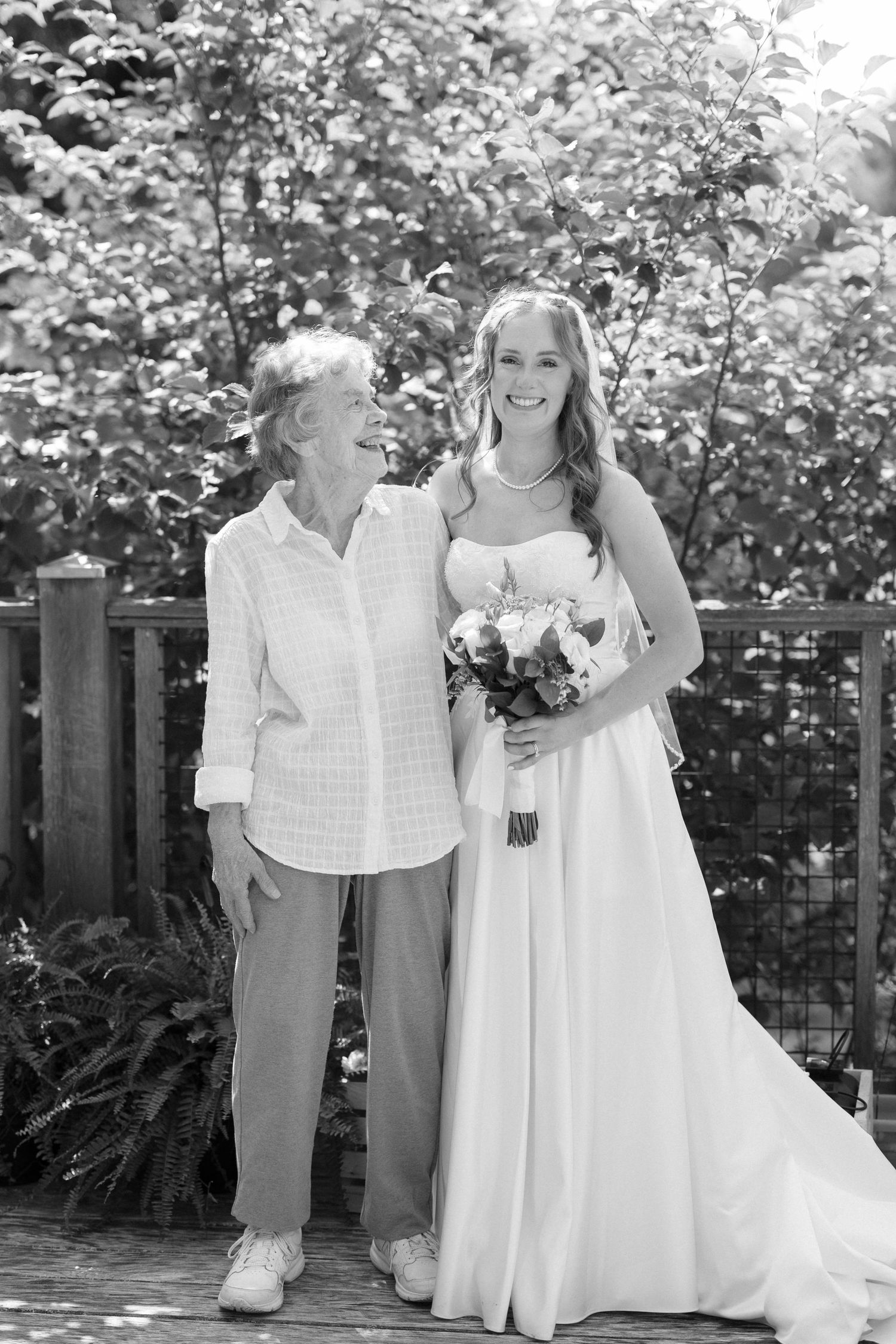 Black and white portrait of bride with elderly woman on wooden deck surrounded by foliage.