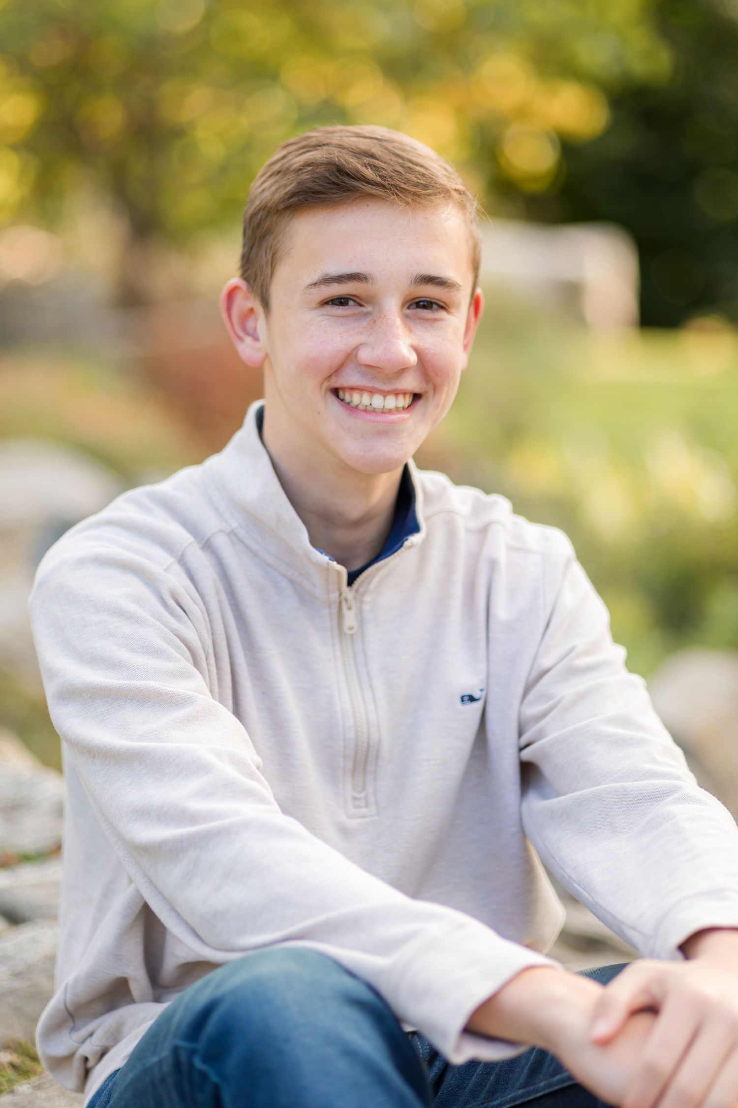 A smiling teenage boy in a light zip-up sweater sitting outdoors among rocks with autumn foliage behind him.