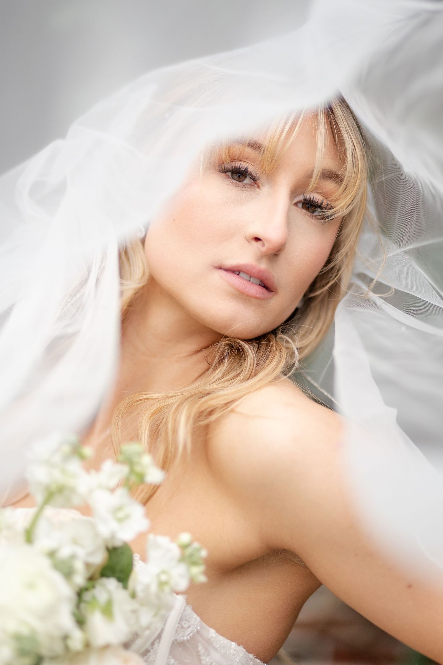 Close-up of blonde bride under flowing white veil holding white floral bouquet.