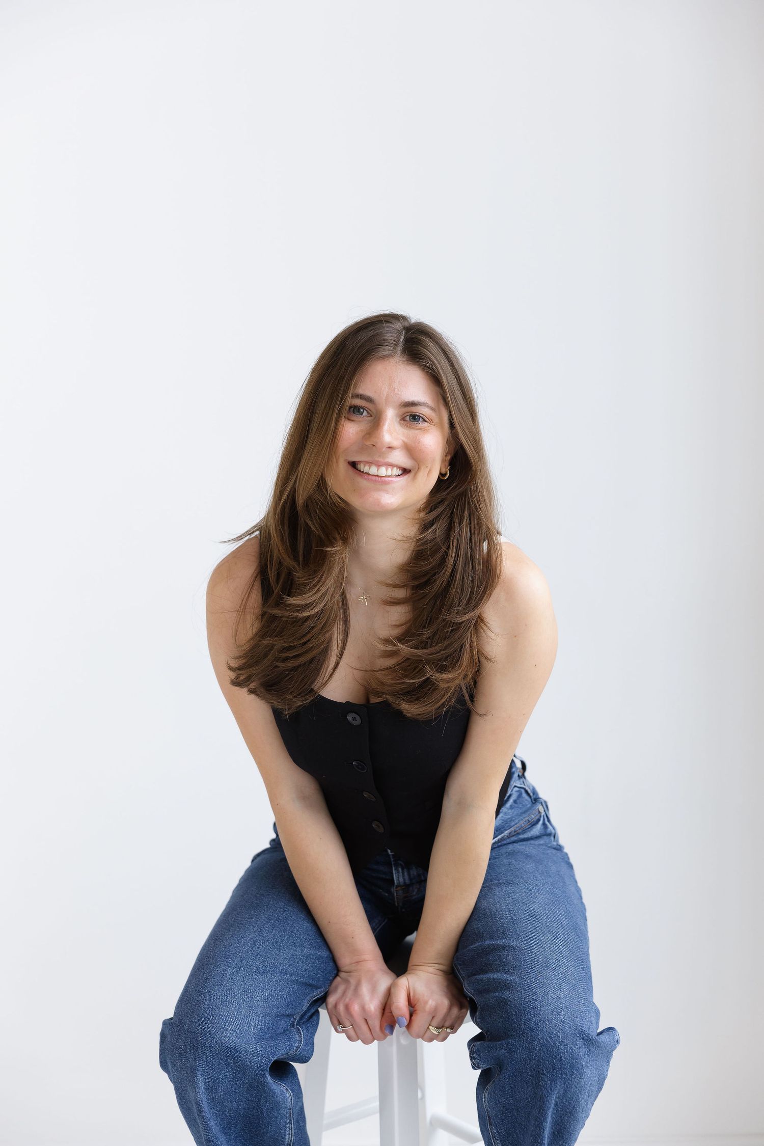 Smiling young woman with long brown hair sitting on stool wearing black top and jeans.