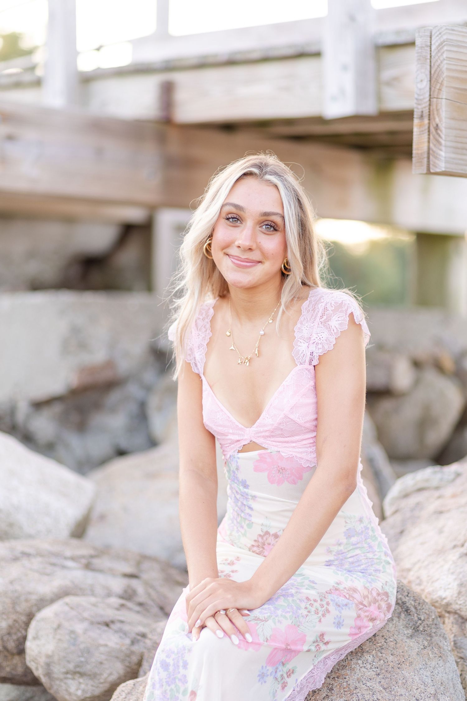Blonde woman in pink floral dress sitting on rocks near a wooden pier, smiling at camera.