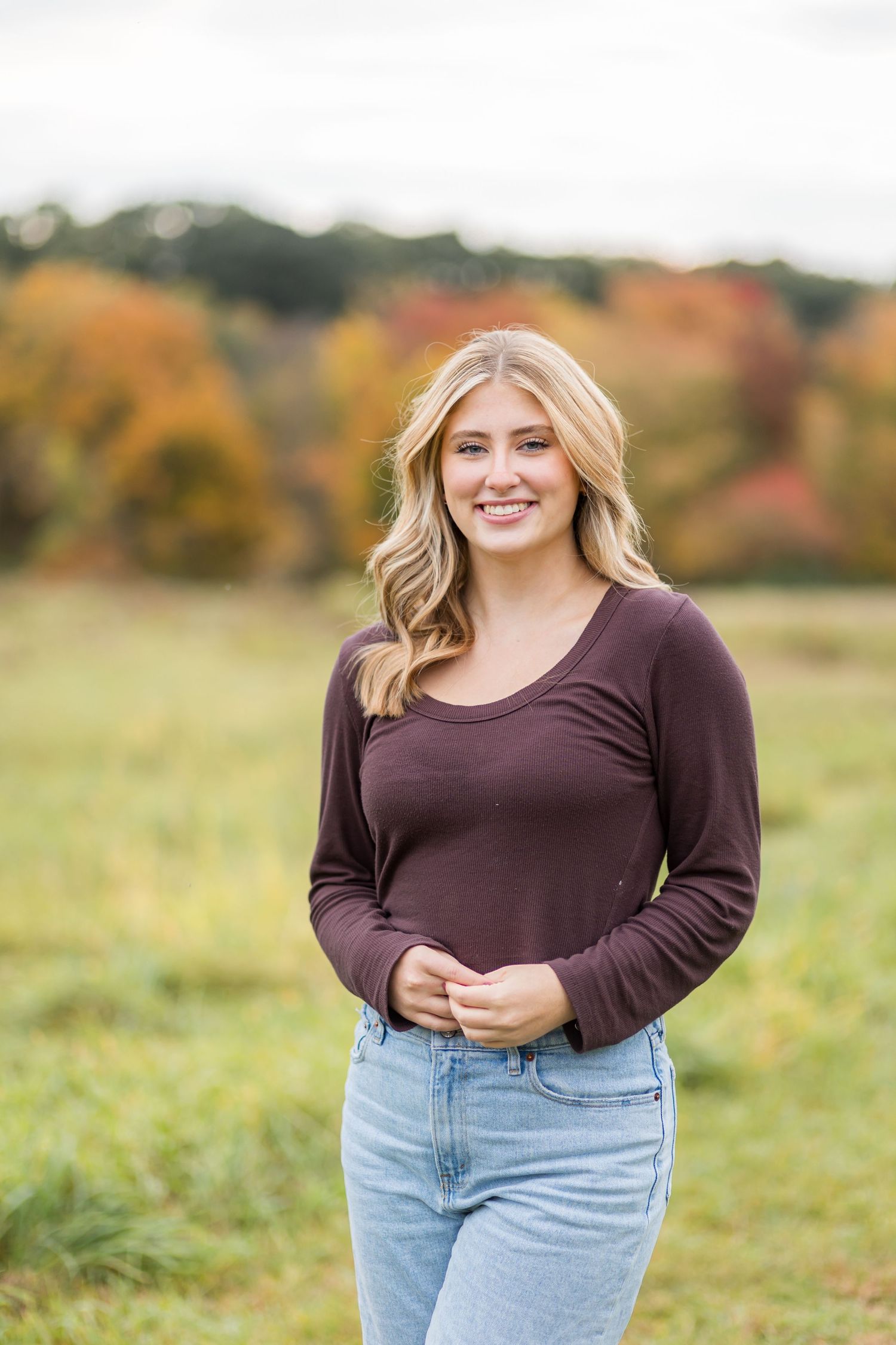 Smiling blonde woman in burgundy long-sleeve top and jeans posing in colorful autumn field.