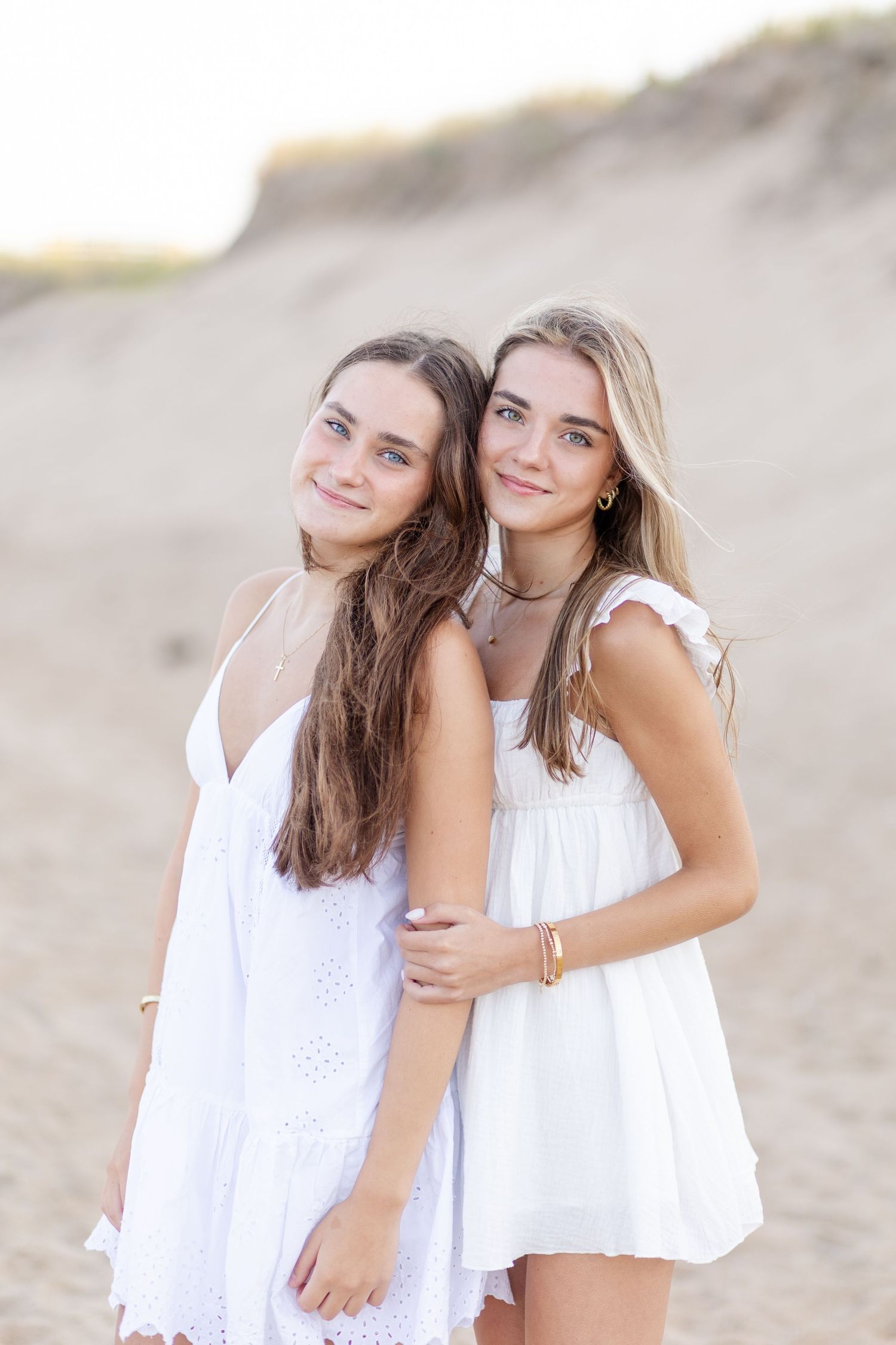 Two teen girls in white dresses embrace on sandy beach dunes at golden hour.