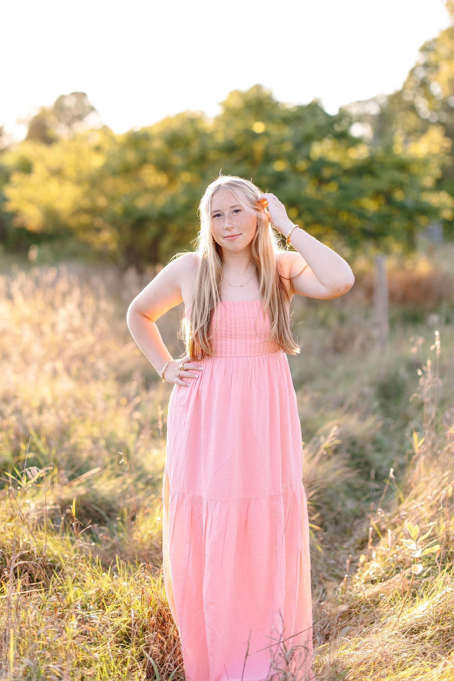 Blonde teen girl in pink maxi dress standing in sunlit field with trees behind her.