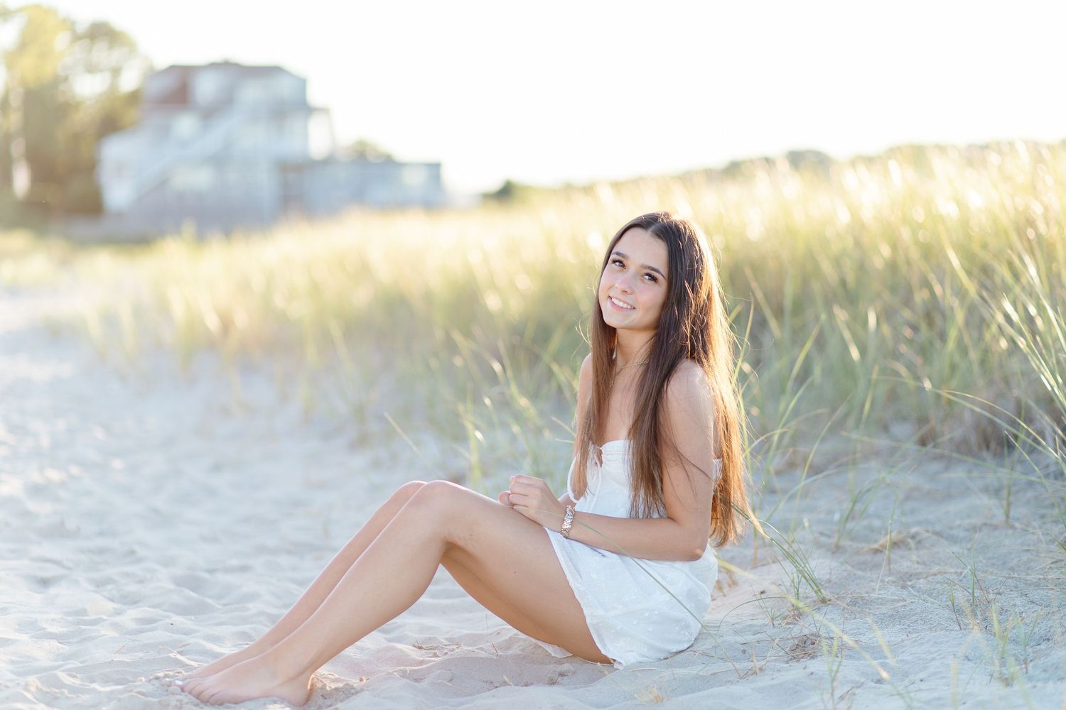 Young woman with long hair sitting on sandy beach path surrounded by golden beach grass at sunset.