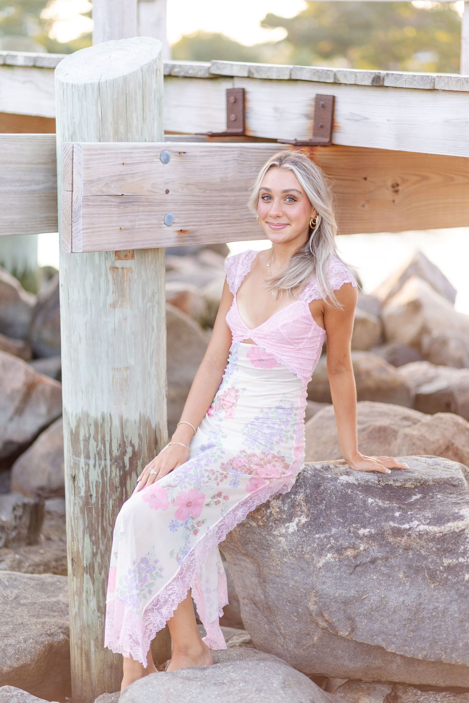 Young blonde woman in pink floral maxi dress sitting on rocks near wooden pier at beach.
