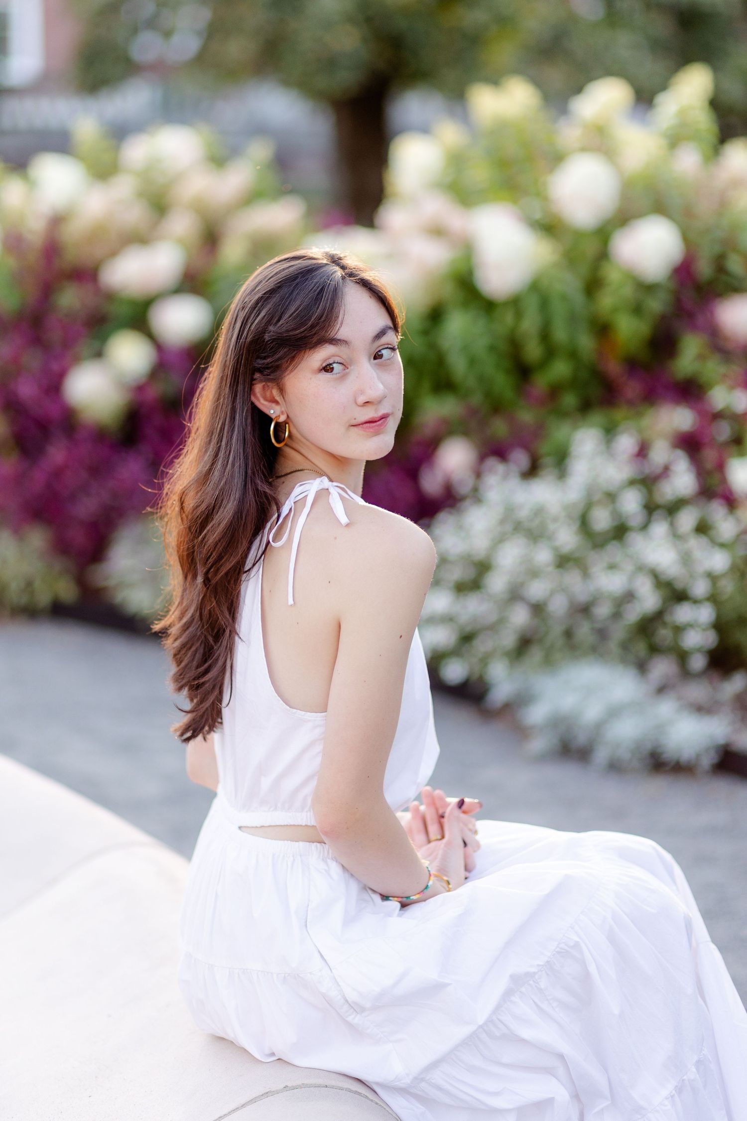 Young Asian woman in white sundress sitting near colorful garden flowers, glancing over shoulder.