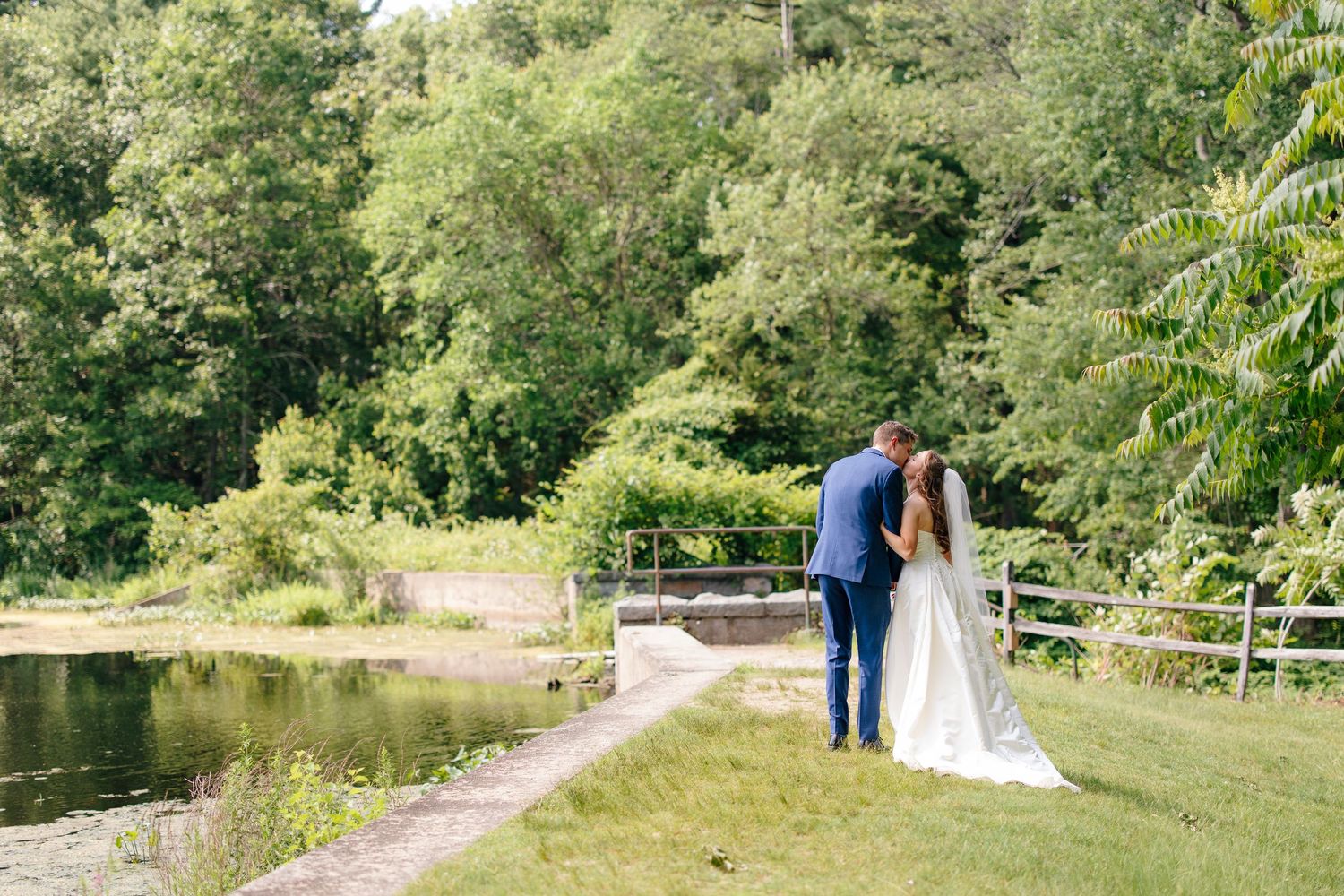 Bride and groom sharing romantic kiss near scenic pond surrounded by lush green landscape.