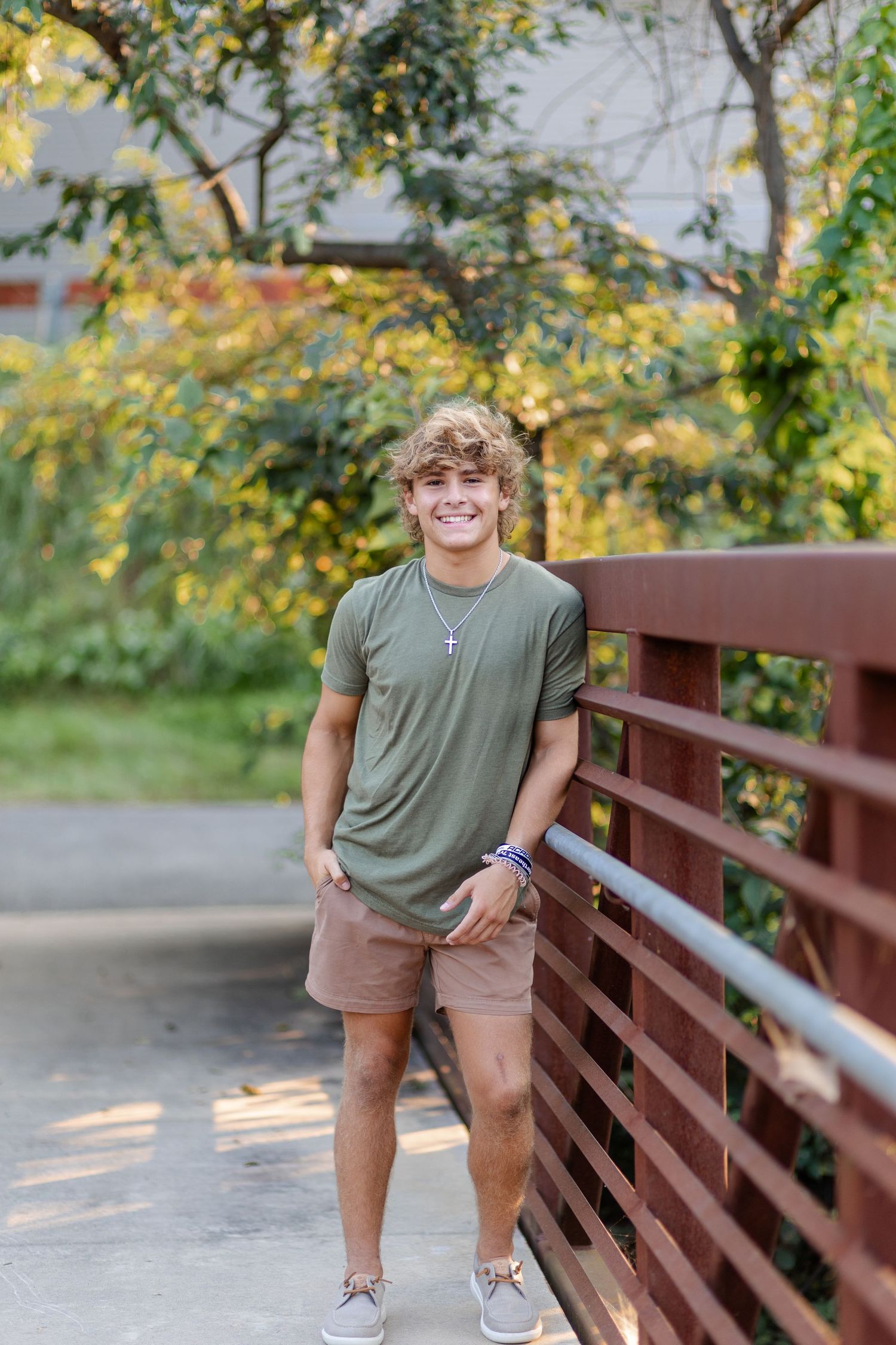 A smiling teenage boy in a green t-shirt and khaki shorts leaning against a wooden bridge railing outdoors.